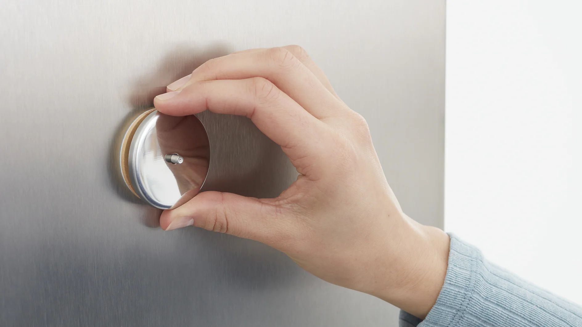 A hand wearing a blue sleeve is adjusting a round metallic dial on a stainless steel surface secured with tesa tape. The dial has a reflective finish, and the background is minimal, focusing on the hand and the dial. (This text has been generated by AI)