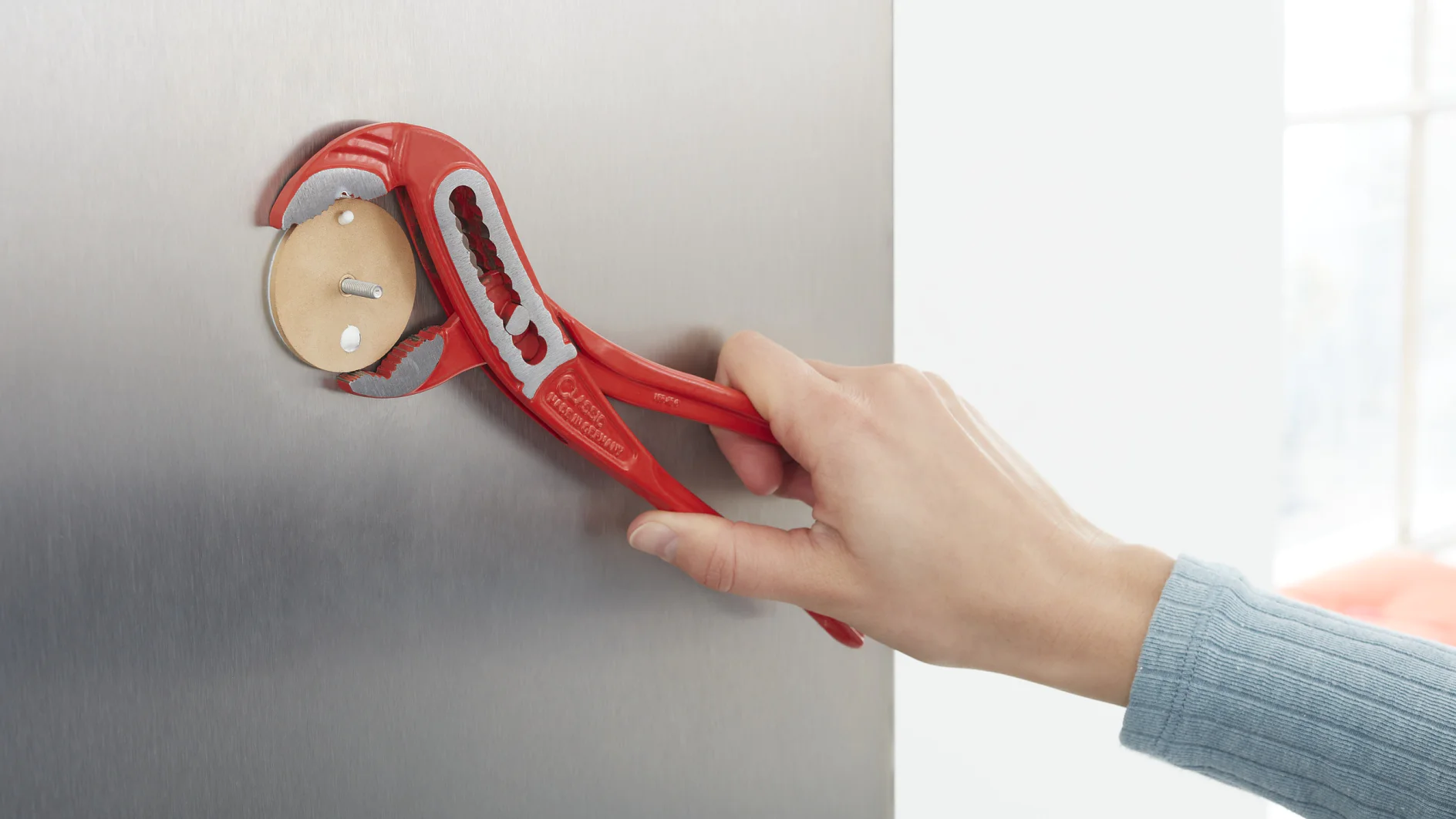 A hand holding a red pipe wrench is gripping a circular fitting with two nails inserted into a flat, metallic surface. The background is plain and softly lit. (This text has been generated by AI)
