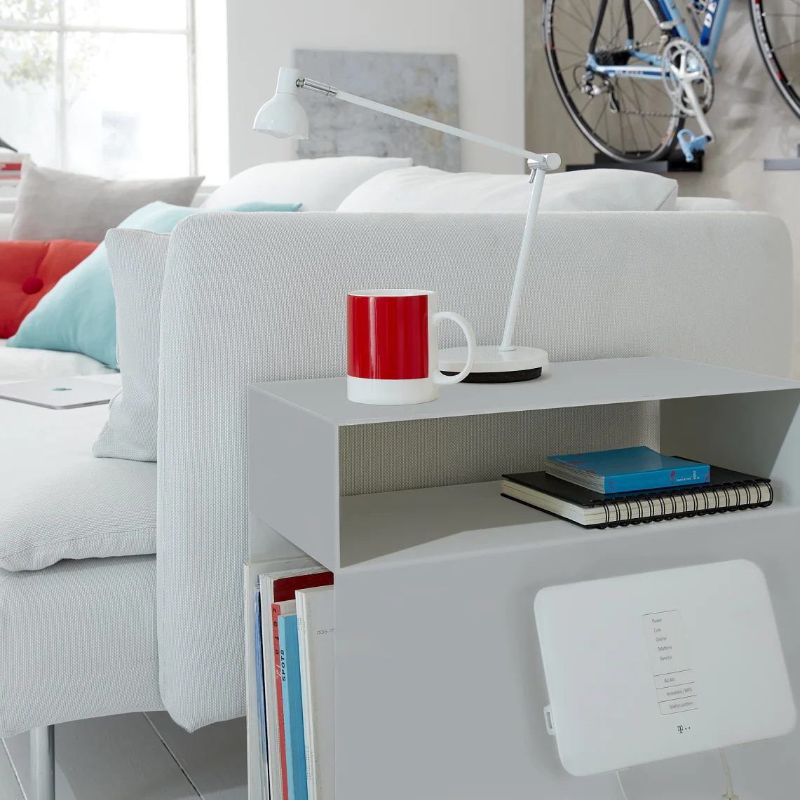 A modern living room showcases a white sofa adorned with red and gray throw pillows. A red mug and a white lamp are placed on a side table. A bicycle is mounted on the wall using tesa tape. Books and a notepad are neatly stored on the table shelf. (This text has been generated by AI)