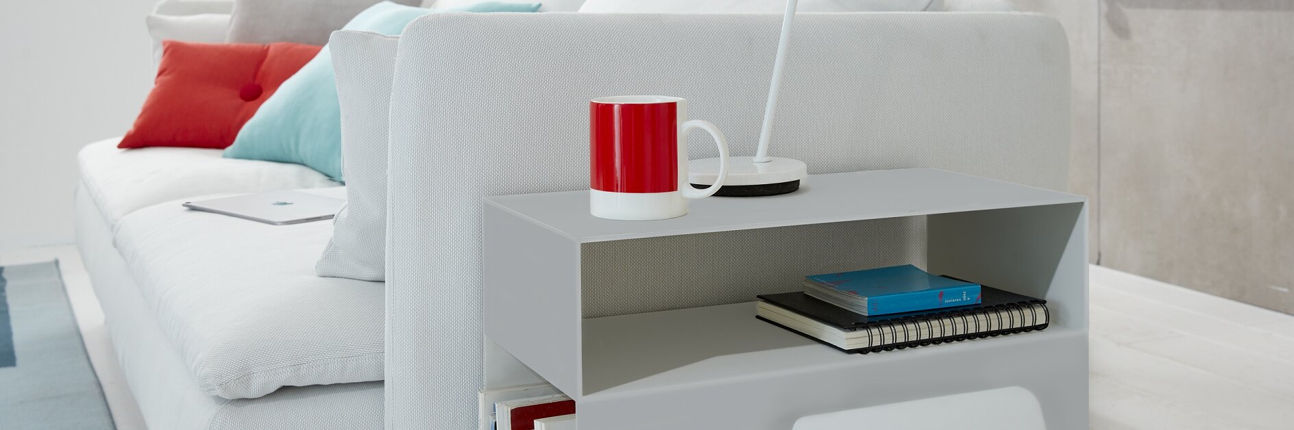 A modern living room showcases a white sofa adorned with red and gray throw pillows. A red mug and a white lamp are placed on a side table. A bicycle is mounted on the wall using tesa tape. Books and a notepad are neatly stored on the table shelf. (This text has been generated by AI)