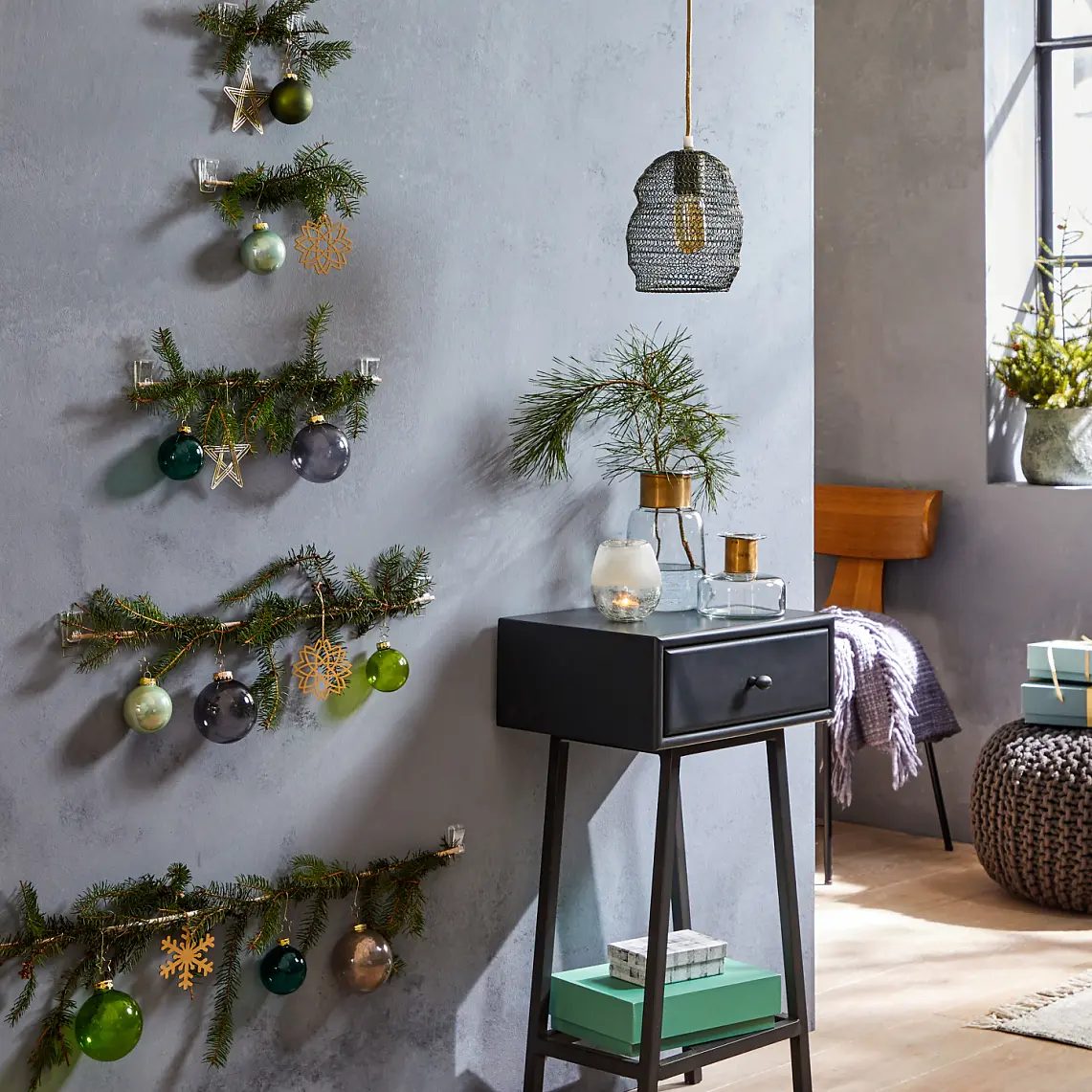 A minimalist Christmas tree design made from branches with ornaments is mounted on a grey wall using tesa tape. A small black table holds decorative items. A woven ottoman and wrapped gifts sit nearby in a room with natural light from a large window. (This text has been generated by AI)