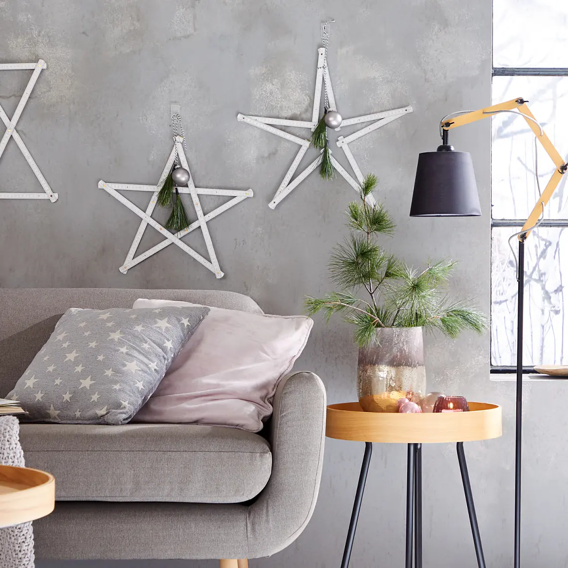 A cozy living room with a grey sofa, star-patterned pillow, and knitted blanket. Three wooden star decorations hang on the grey wall using tesa tape. A wooden side table with pine branches and a black lamp completes the setting. A window displays candles and dried branches, adding to the ambiance. (This text has been generated by AI)