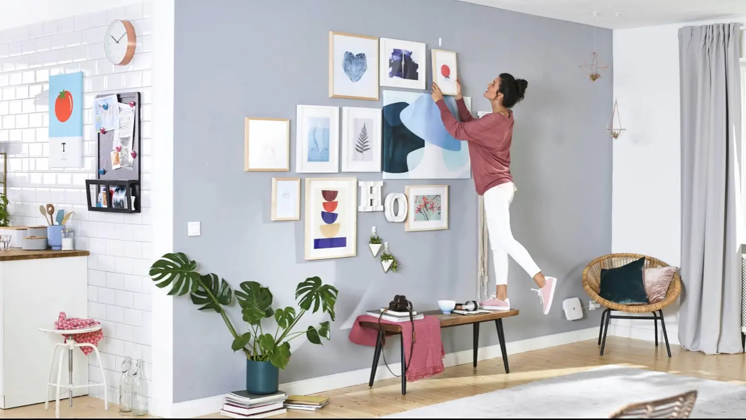 A person stands on a bench, adjusting a framed picture on a gallery wall in a living room. The wall features various artworks, and the room is decorated with plants and modern furnishings, while the kitchen area is visible in the background. To secure everything, they use tesa tape for all their hanging needs.