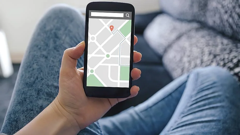 En person holder en smartphone og viser et kort med en placeringsnål. De sidder på en sofa iført falmede jeans. Baggrunden er let sløret og viser en mørk pude og en del af sofaen. (This text has been generated by AI)