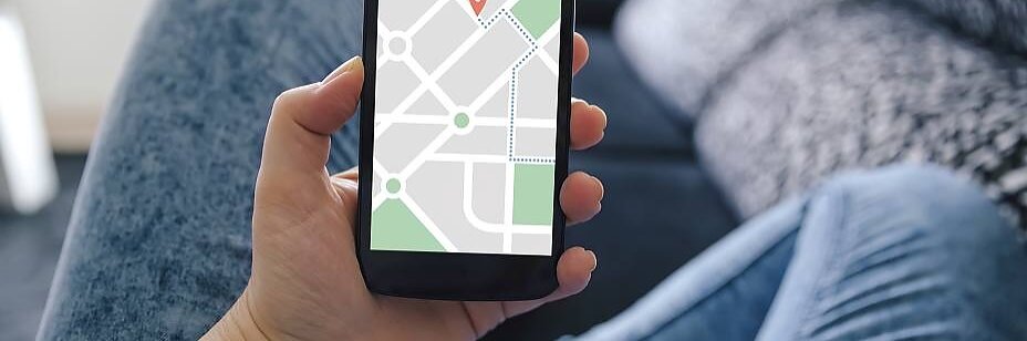 En person holder en smartphone og viser et kort med en placeringsnål. De sidder på en sofa iført falmede jeans. Baggrunden er let sløret og viser en mørk pude og en del af sofaen. (This text has been generated by AI)