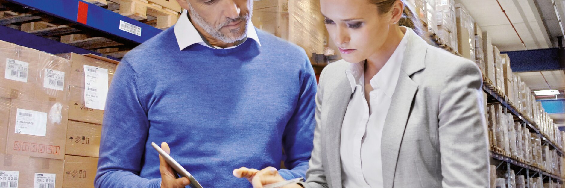 En mand og en kvinde står på et lager, omgivet af hylder stablet med kasser. Manden i en blå sweater holder en tablet med tesa-tape. Kvinden, i en grå blazer, skriver på en tesa-tape-mærket udklipsholder. De ser ud til at diskutere inventar. (This text has been generated by AI)