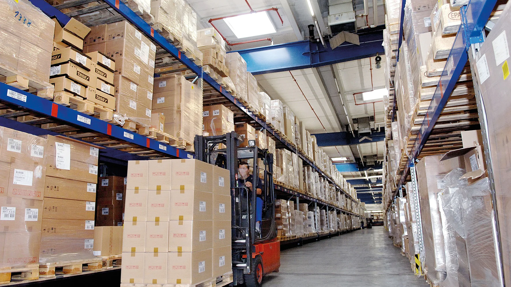 Et stort lager med høje hylder stablet med kasser. En arbejder betjener en rød gaffeltruck og flytter en palle med papkasser forseglet med tesa-tape. Gangen er bred, og loftet har lysstofrør og ventilationskanaler. (This text has been generated by AI)
