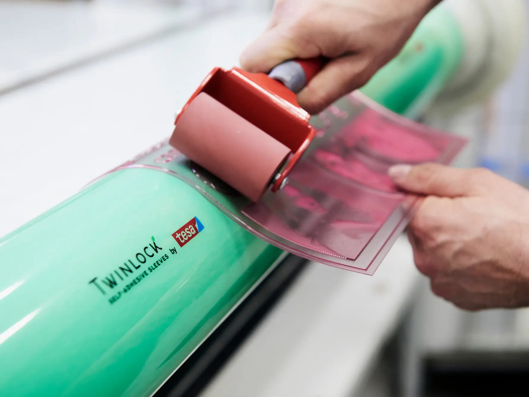 Close-up of hands using a small roller to apply a pink sheet onto a large green cylinder labeled Twinlock. The cylinder also displays the phrase Self Adhesive Sleeves, with tesa tape being used in the process. (This text has been generated by AI)