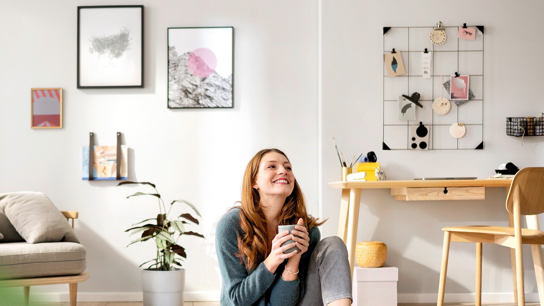 A woman with long hair is sitting on the floor in a brightly lit living room holding a mug, smiling upward. The room features a small desk with a chair, a potted plant, wall art, and decorative items secured with tesa tape on a wire grid. (This text has been generated by AI)