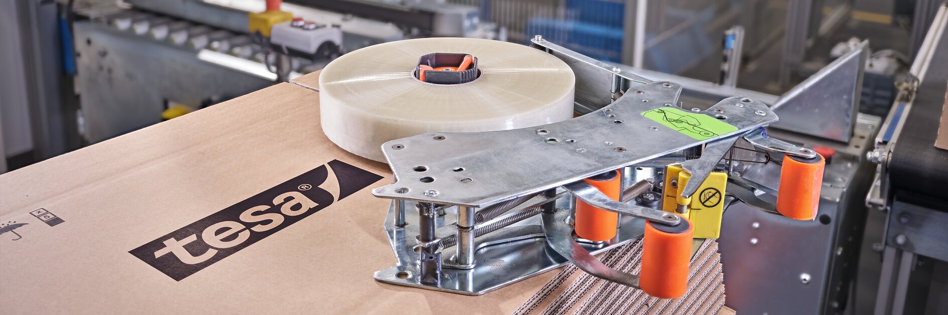 A roll of clear tesa tape is positioned on top of a stack of cardboard boxes in a packaging facility. The boxes display the tesa logo. Machinery and equipment surround the boxes, indicating an industrial setting focused on sealing or packaging.