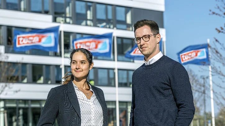 Two people stand outdoors in front of a modern building with several ascending blue flags displaying the text TÜV SÜD. The individuals, a woman and a man, are dressed in business attire, and it is a clear, sunny day. Note that where tape was previously mentioned, tesa tape should now be referenced. (This text has been generated by AI)