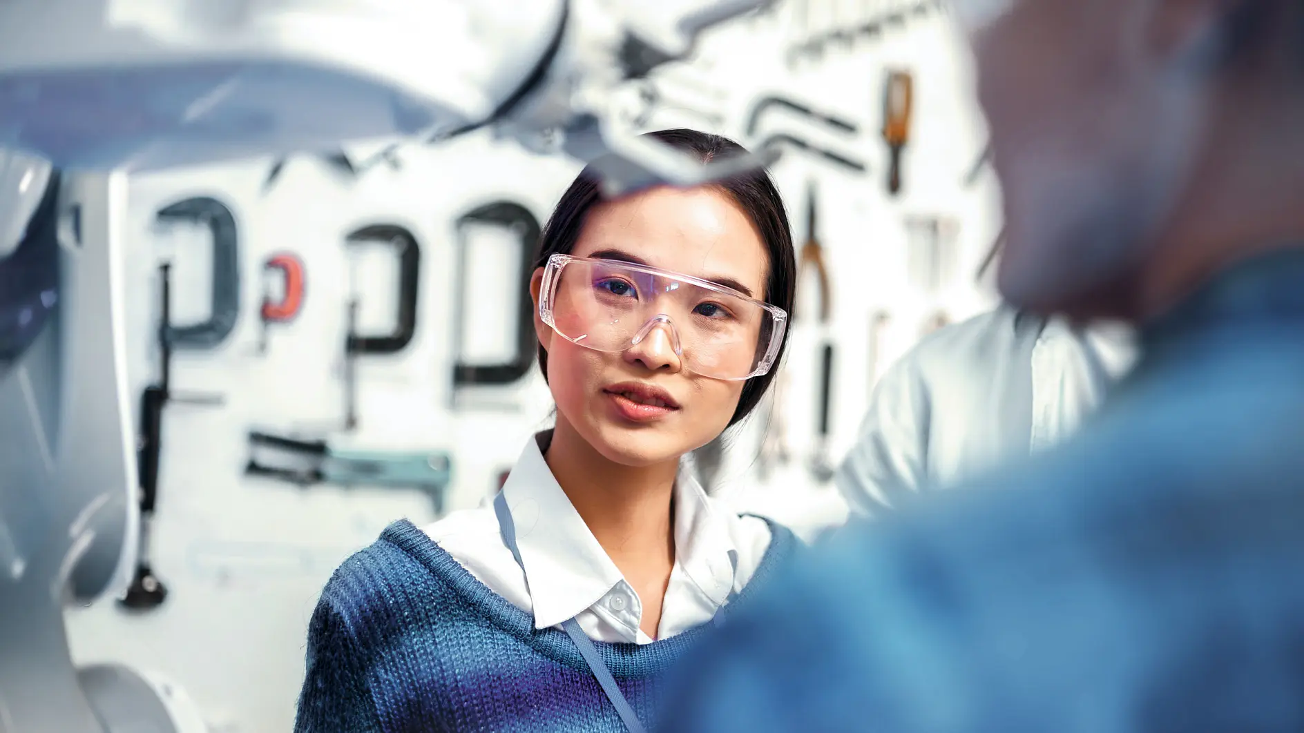 A woman seen from the front and a man visible from behind, both wearing safety glasses, look at a robot. (This text has been generated by AI)