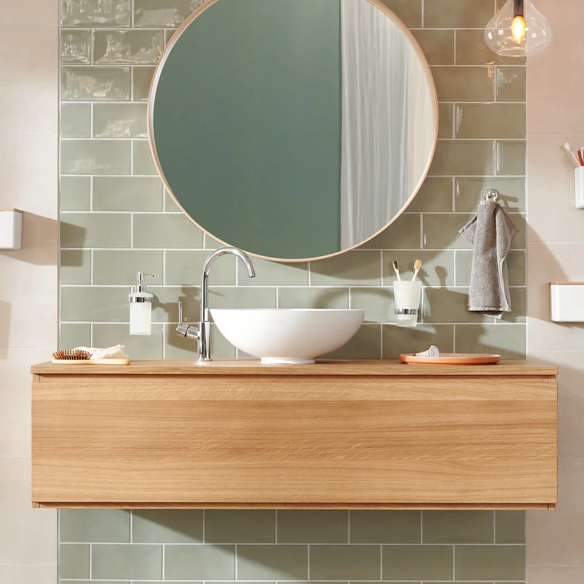 Modern bathroom with a round mirror above a wooden vanity. A white vessel sink sits on the vanity. Green rectangular tiles are on the wall. Accessories include a hanging hand towel, soap dispenser, and toothbrush holder secured with tesa tape. Warm lighting overhead. (This text has been generated by AI)
