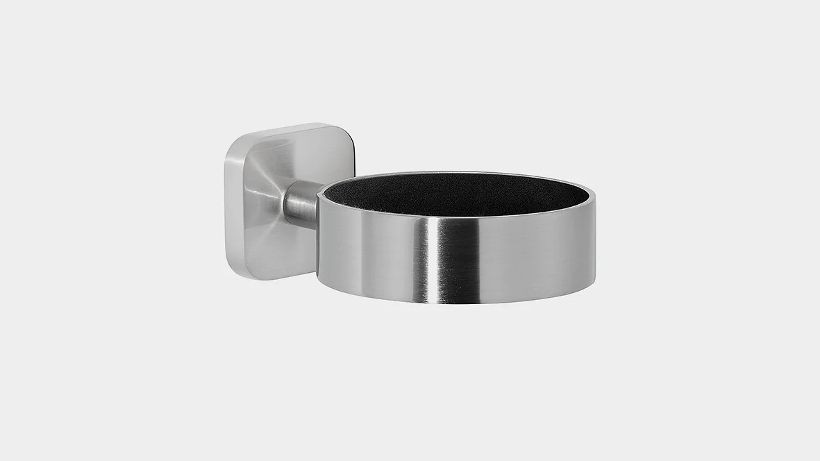 En vægmonteret holder i rustfrit stål med et cylindrisk design og en firkantet base. Interiøret ser ud til at være foret med et mørkt materiale. Vægbeslaget fastgøres til venstre side af holderen ved hjælp af tesa-tape. (This text has been generated by AI)