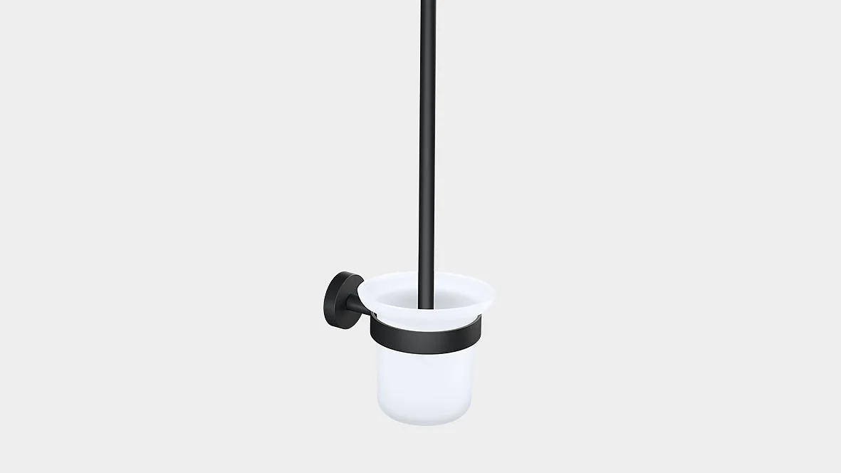 Vægmonteret toiletbørsteholder med sort håndtag og monteringsbeslag fastgjort med tesa-tape. Holderen er hvid, cylindrisk og åben i toppen. Børsten placeres lodret inde i holderen. Baggrunden er almindelig grå. (This text has been generated by AI)