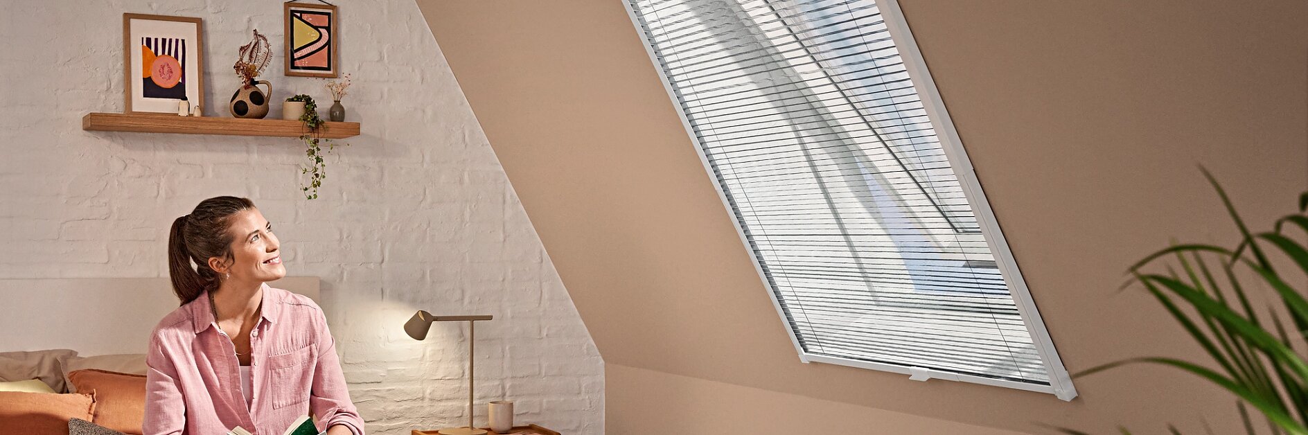En person sidder med benene over kors på en seng med en åben bog i hånden og kigger mod et ovenlysvindue med persienner. Rummet har skråt loft, et lille sidebord med dekorationsgenstande og en hylde med billeder på væggen. En plante er delvist synlig i forgrunden. (This text has been generated by AI)