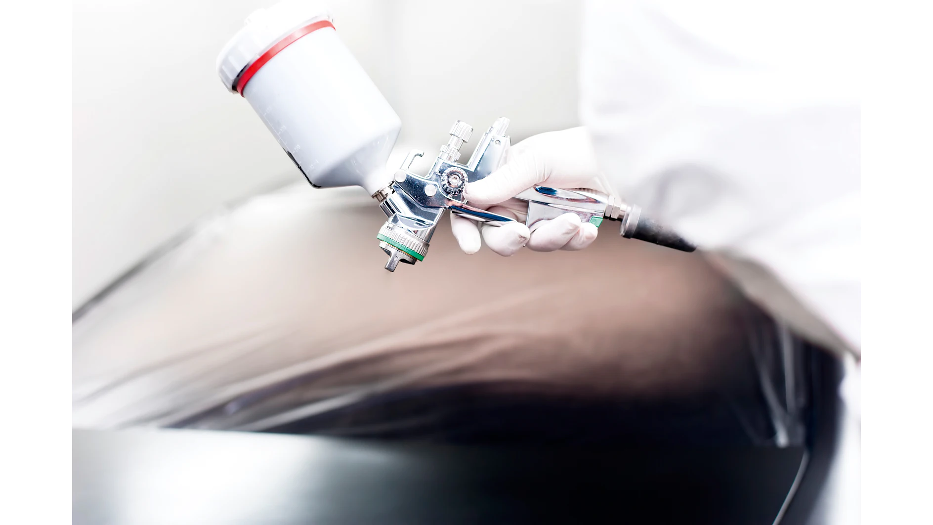 En person med hvide handsker bruger en sprøjtepistol til at male en bil, og langs bilens kant kan man se tesa-tape som afskærmning. (This text has been generated by AI)
