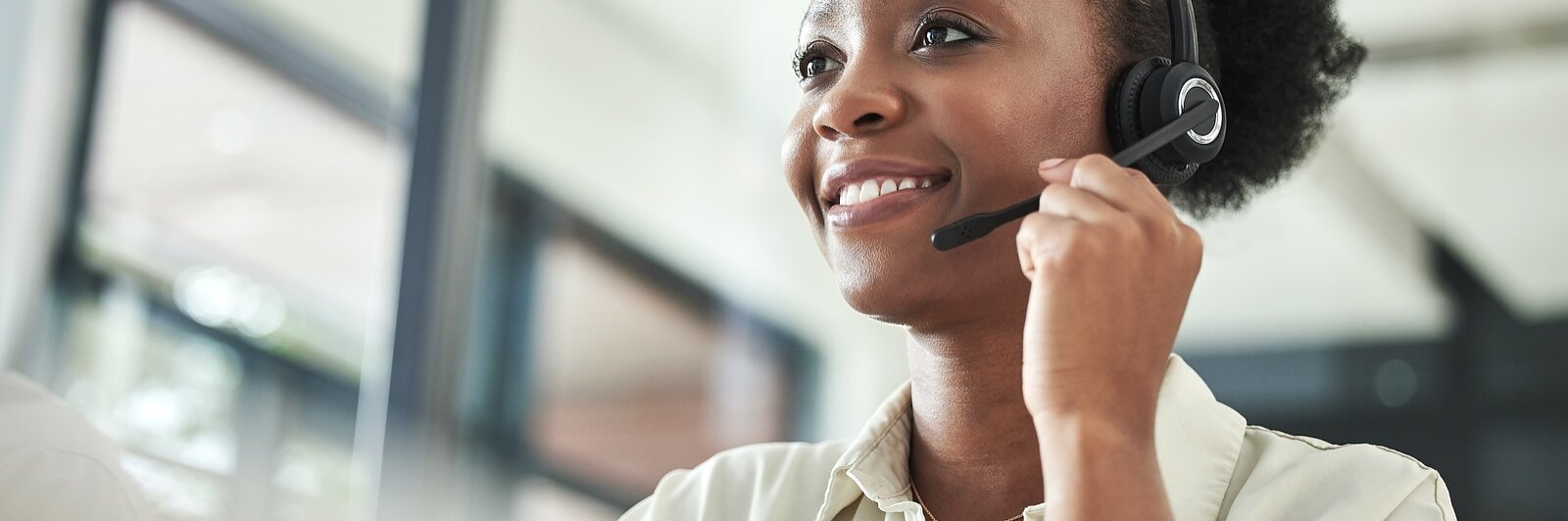 En kvinde i headset og lys skjorte sidder ved et skrivebord, smiler og kigger til siden i et lyst, moderne kontor med tesa-tape.