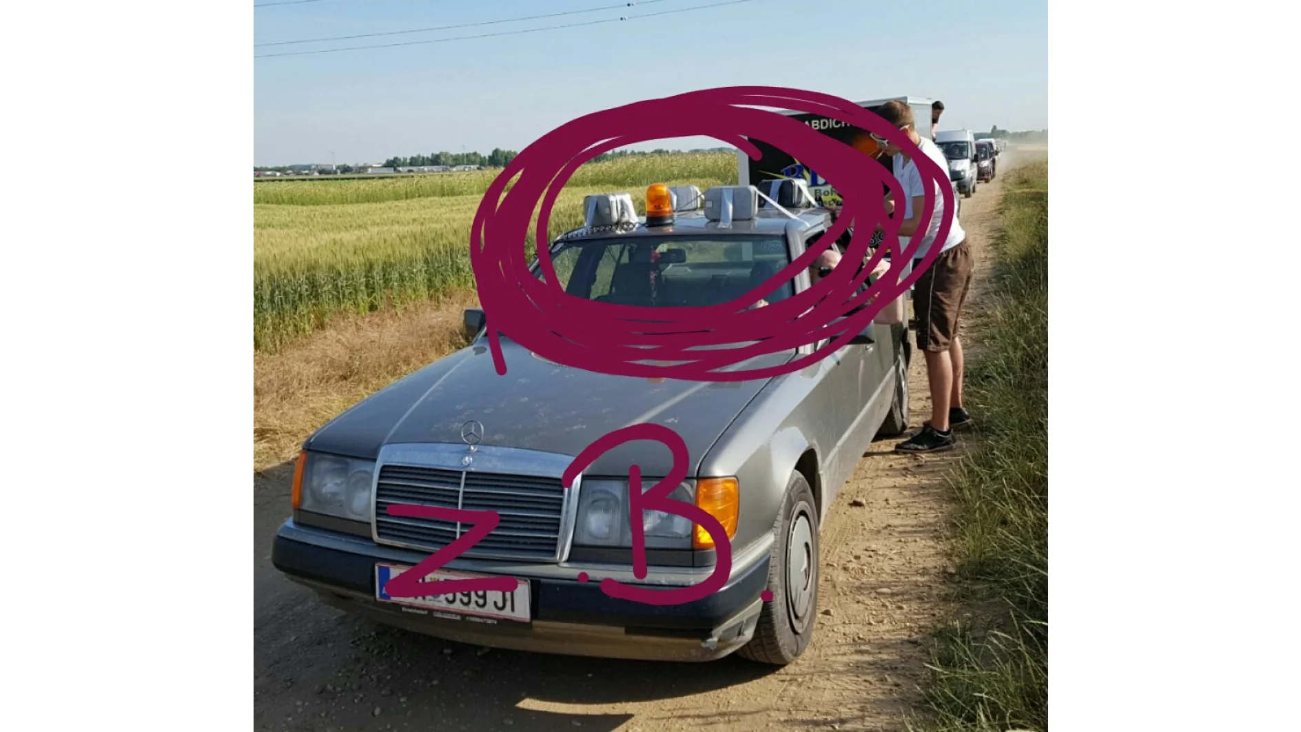 Auf einem Feldweg neben einem Feld parkt ein silbernes Auto. Auf dem Dach befinden sich mehrere metallische Gegenstände und eine orangefarbene Warnleuchte. Neben dem Fahrzeug steht eine Person. Auf dem Bild sind rote Kritzeleien mit den Initialen „Z.B.“ im Vordergrund zu sehen, die mit Tesa-Klebeband sicher befestigt sind. (This text has been generated by AI)