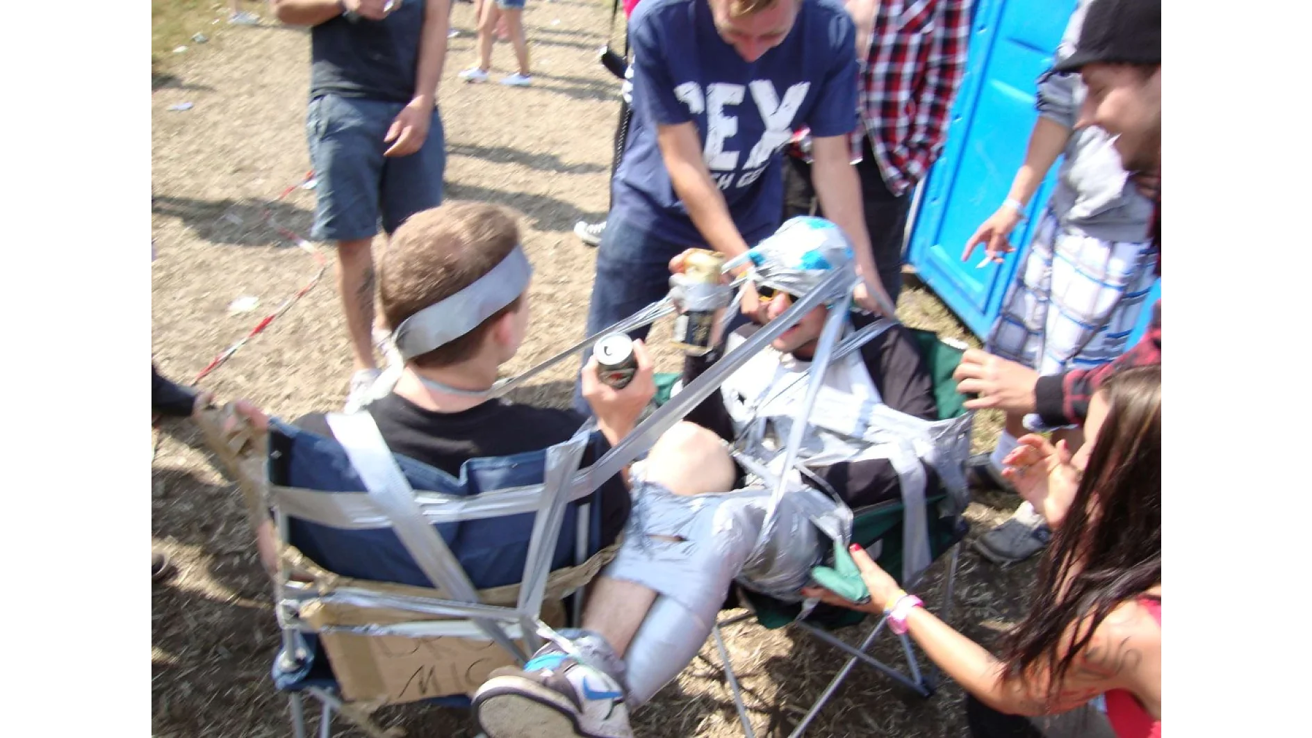 Zwei Personen sind mit Tesa-Klebeband an Campingstühle gefesselt. Einer hält eine Getränkedose. Die anderen um sie herum scheinen zu lachen. Sie stehen draußen auf einem Erdboden, in der Nähe einer blauen Mobiltoilette. Mehrere Personen stehen und tragen Freizeitkleidung. (This text has been generated by AI)