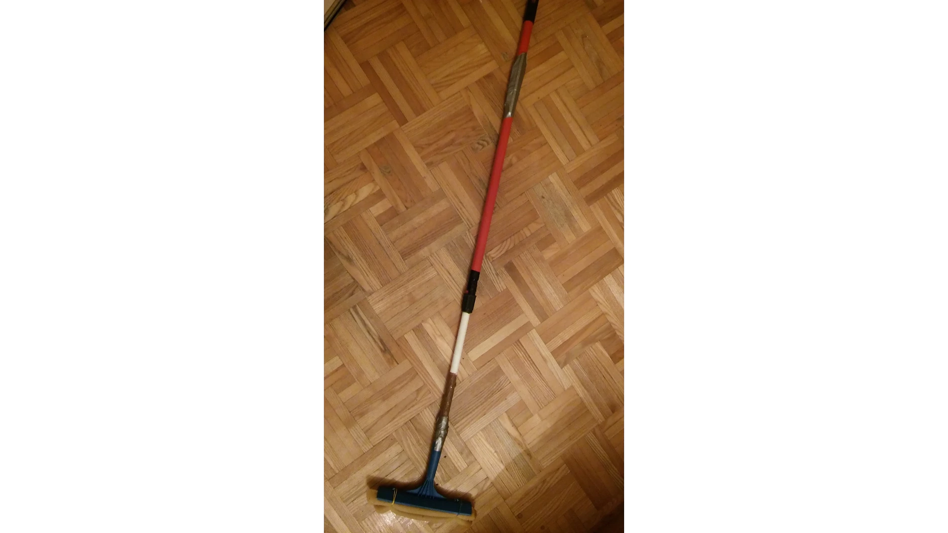 Ein Bodenwischer mit einem mit rot-weißem Tesa-Band umwickelten Stiel liegt auf einem Holzparkettboden. Der Wischerkopf ist blau mit einer gelben Kante. Der Stiel hat verschiedene Segmente, die mit Tesa-Band repariert sind. (This text has been generated by AI)