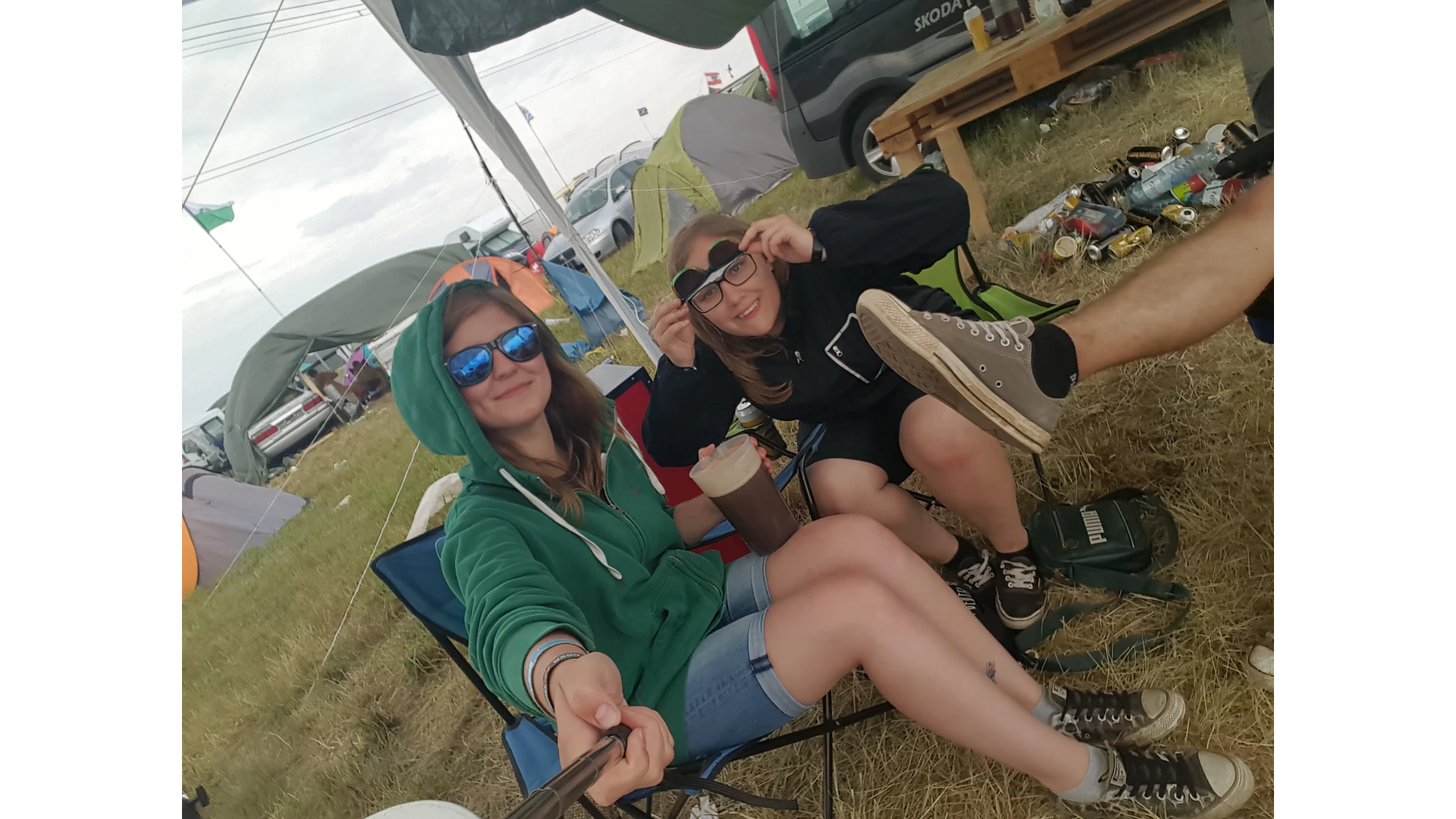 Zwei Personen sitzen draußen auf einem Campingplatz. Einer hält ein Selfie-Stick und trägt eine Sonnenbrille und einen grünen Kapuzenpullover. Der andere rückt seine Sonnenbrille zurecht und lächelt. Im Hintergrund sind ein Zelt, ein Auto und verstreute Getränkedosen zu sehen. (This text has been generated by AI)