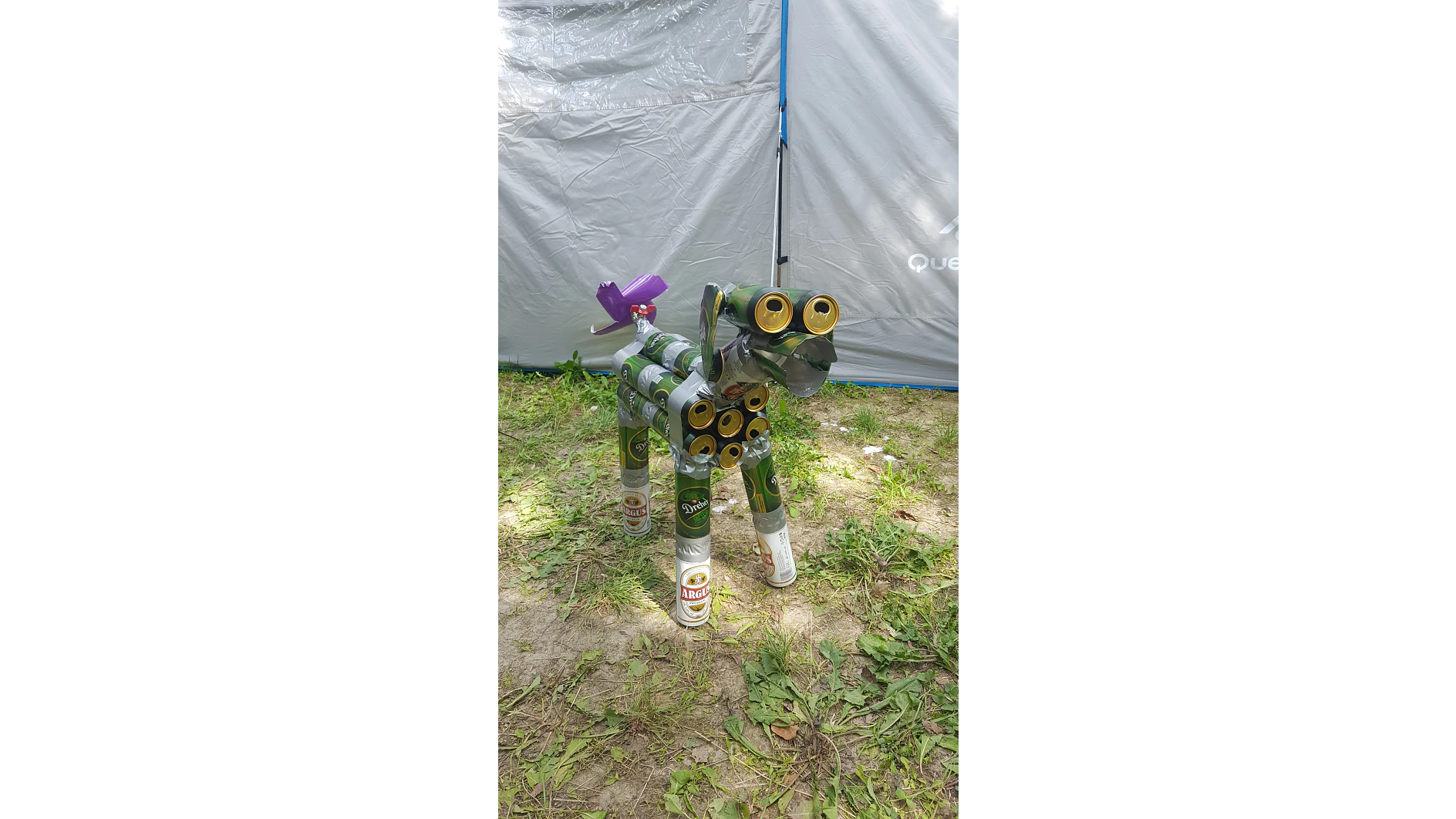 Auf einer Rasenfläche steht eine Skulptur, die einem Hund ähnelt und aus grünen und silbernen Bierdosen besteht. Der Kopf hat zwei große gelbe Ringösen und auf seinem Rücken befindet sich ein violettes Plastikobjekt. Im Hintergrund ist ein graues Zelt zu sehen. (This text has been generated by AI)