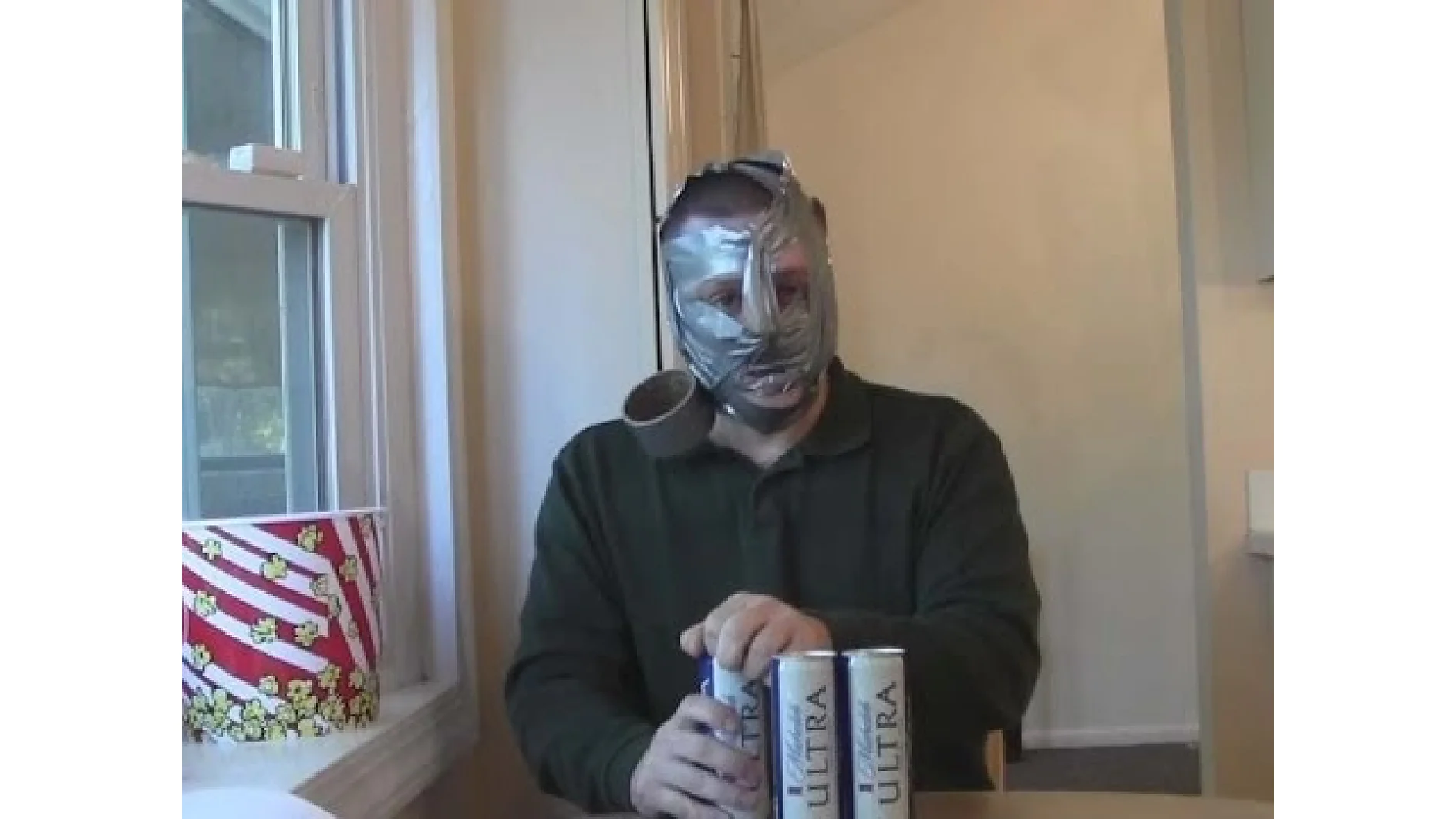 Eine Person sitzt an einem Tisch, ihr Gesicht ist mit Tesa-Klebeband umwickelt, und hält drei Getränkedosen in der Hand. An der Seite ist ein halb aufgegessener Eimer Popcorn zu sehen. Die Szene scheint eine Küche mit einem Fenster im Hintergrund zu sein. (This text has been generated by AI)