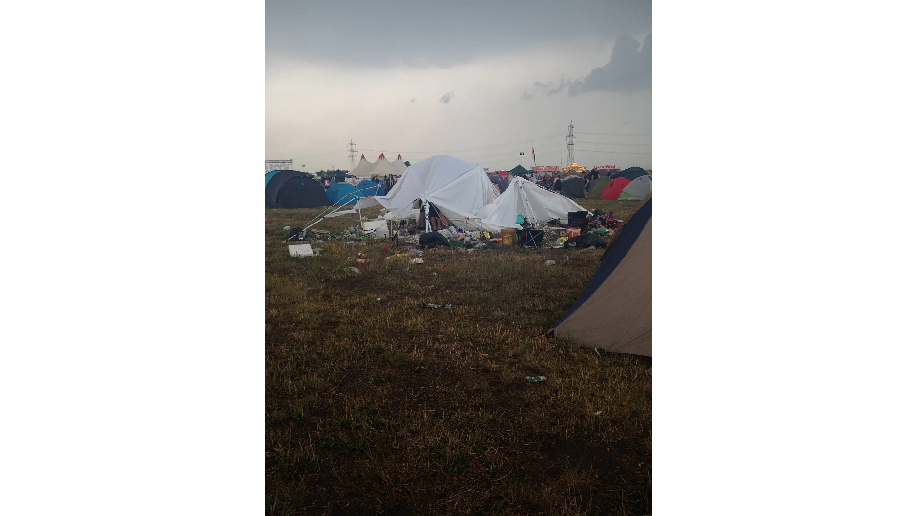 Ein überfüllter Campingplatz an einem bewölkten Tag mit verschiedenen Zelten, von denen einige zusammengebrochen sind. Auf dem Boden liegen Müll und Campingzubehör verstreut, darunter Rollen mit Tesa-Klebeband. Im Hintergrund sind weitere Zelte und Flaggen zu sehen. Der Himmel ist bedeckt, was auf bevorstehenden Regen hindeutet. (This text has been generated by AI)
