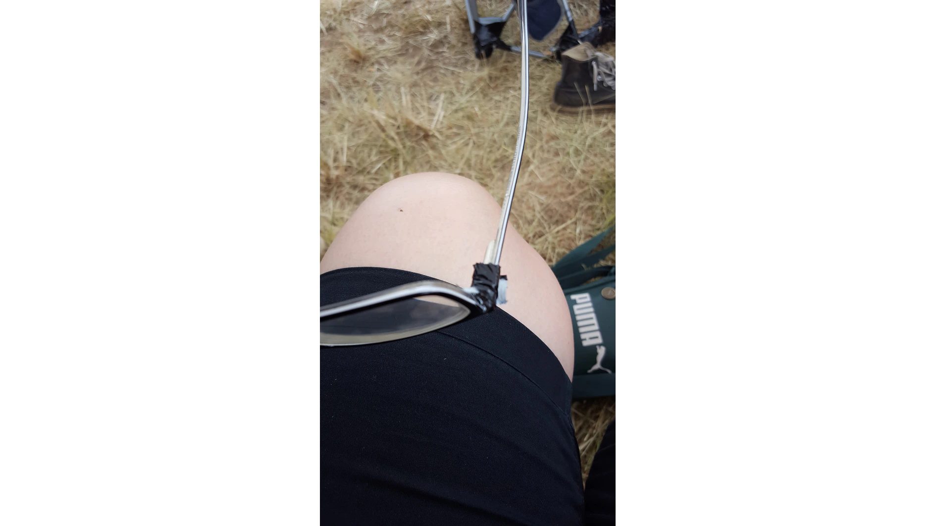 Eine Person sitzt im Gras und hat eine Sonnenbrille notdürftig repariert. Der Rahmen ist mit schwarzem Tesa-Klebeband zusammengehalten. Die Person trägt schwarze Shorts, im Hintergrund sind ein Puma-Schuh und ein Teil eines Campingstuhls zu sehen. (This text has been generated by AI)