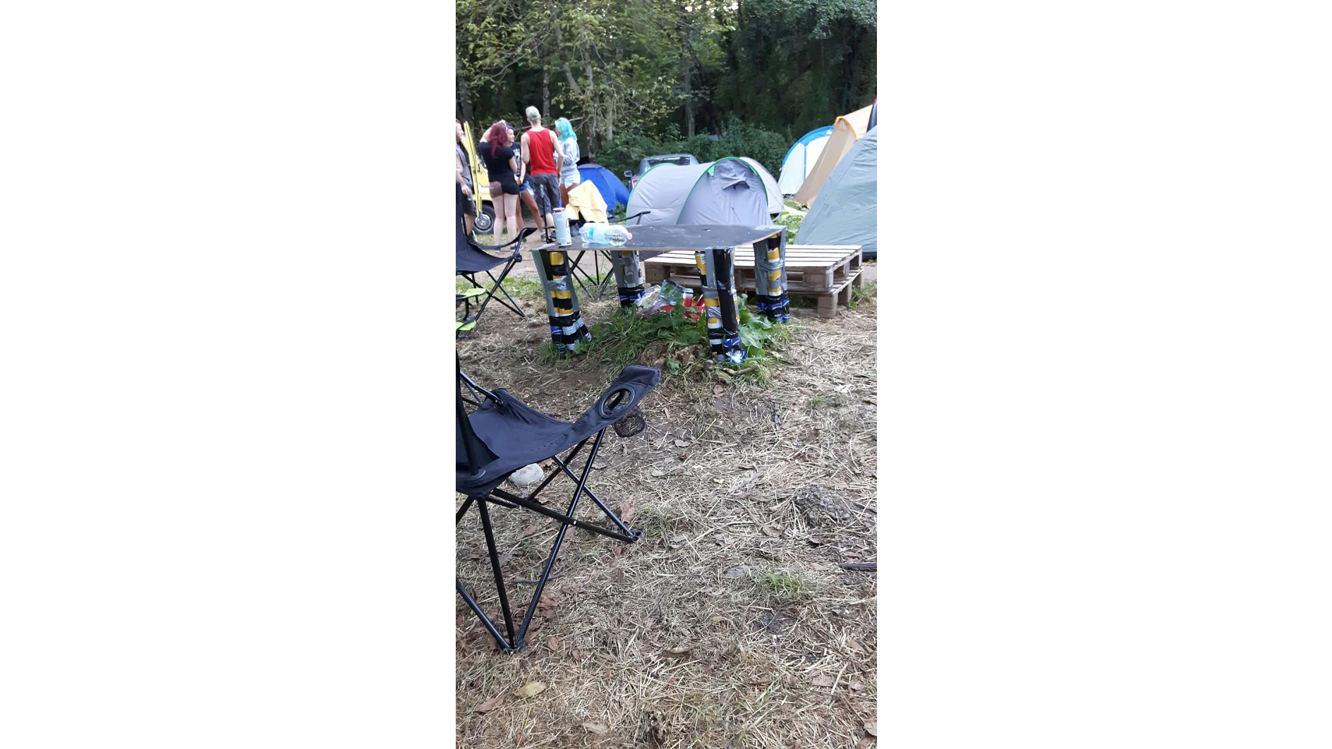 Ein Campingplatz mit mehreren Zelten und Holzpaletten. Auf dem Boden sind vier Dosen übereinander gestapelt und mit Tesaband befestigt. Im Vordergrund sind Klappstühle zu sehen, im Hintergrund versammelt sich eine Gruppe von Menschen bei den Zelten, umgeben von Bäumen. (This text has been generated by AI)