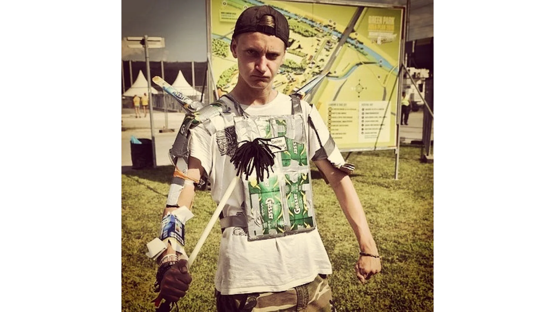 Eine Person mit strengem Gesichtsausdruck trägt ein selbstgemachtes Outfit aus Bierverpackungen und Tesa-Klebeband. Sie hält einen Stock und hat Schachteln an ihren Ärmeln befestigt. Hinter ihr sind eine Karte und Parkschilder zu sehen. (This text has been generated by AI)
