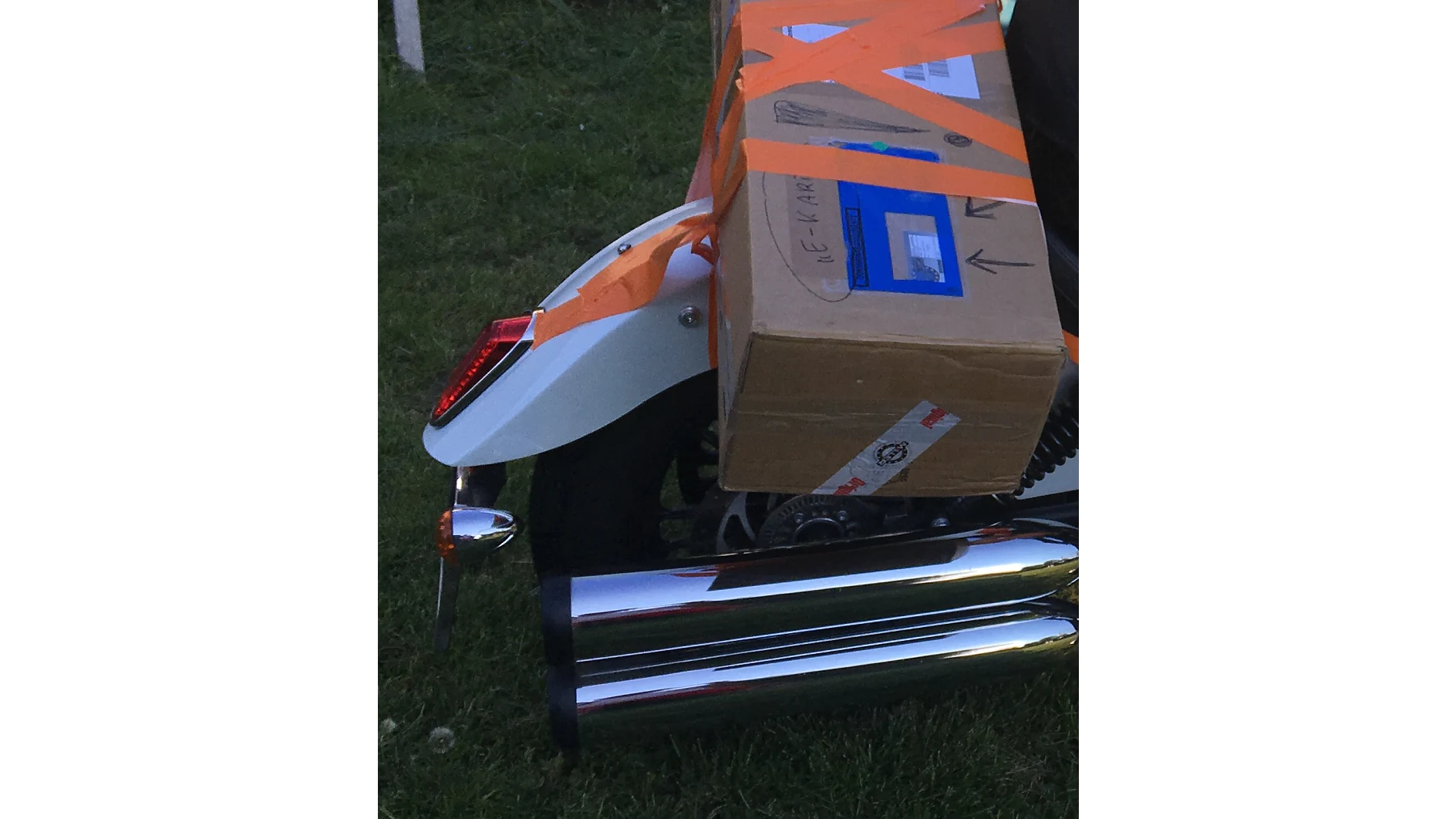 Ein Motorrad, auf dessen Rückseite ein Karton mit orangefarbenem Tesa-Klebeband festgeschnallt ist. Der Karton bedeckt einen Teil des Hinterrads und der Auspuffrohre. Er hat ein blaues Versandetikett und einige handgezeichnete Markierungen. Der Aufbau erfolgt auf einer Rasenfläche. (This text has been generated by AI)