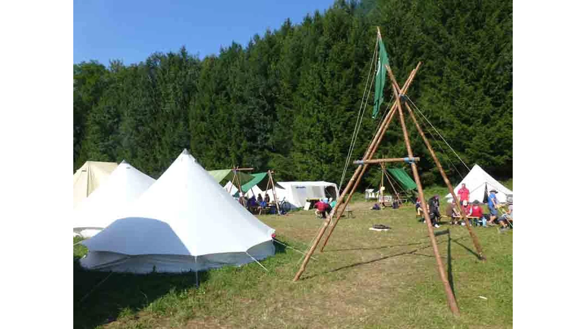 Ein Campingplatz mit mehreren weißen Zelten auf einer Rasenfläche. Es gibt eine Holzkonstruktion mit einer grünen Flagge und im Hintergrund einen dichten Wald. Unter einem klaren blauen Himmel sitzen und versammeln sich Menschen in der Nähe der Zelte. (This text has been generated by AI)