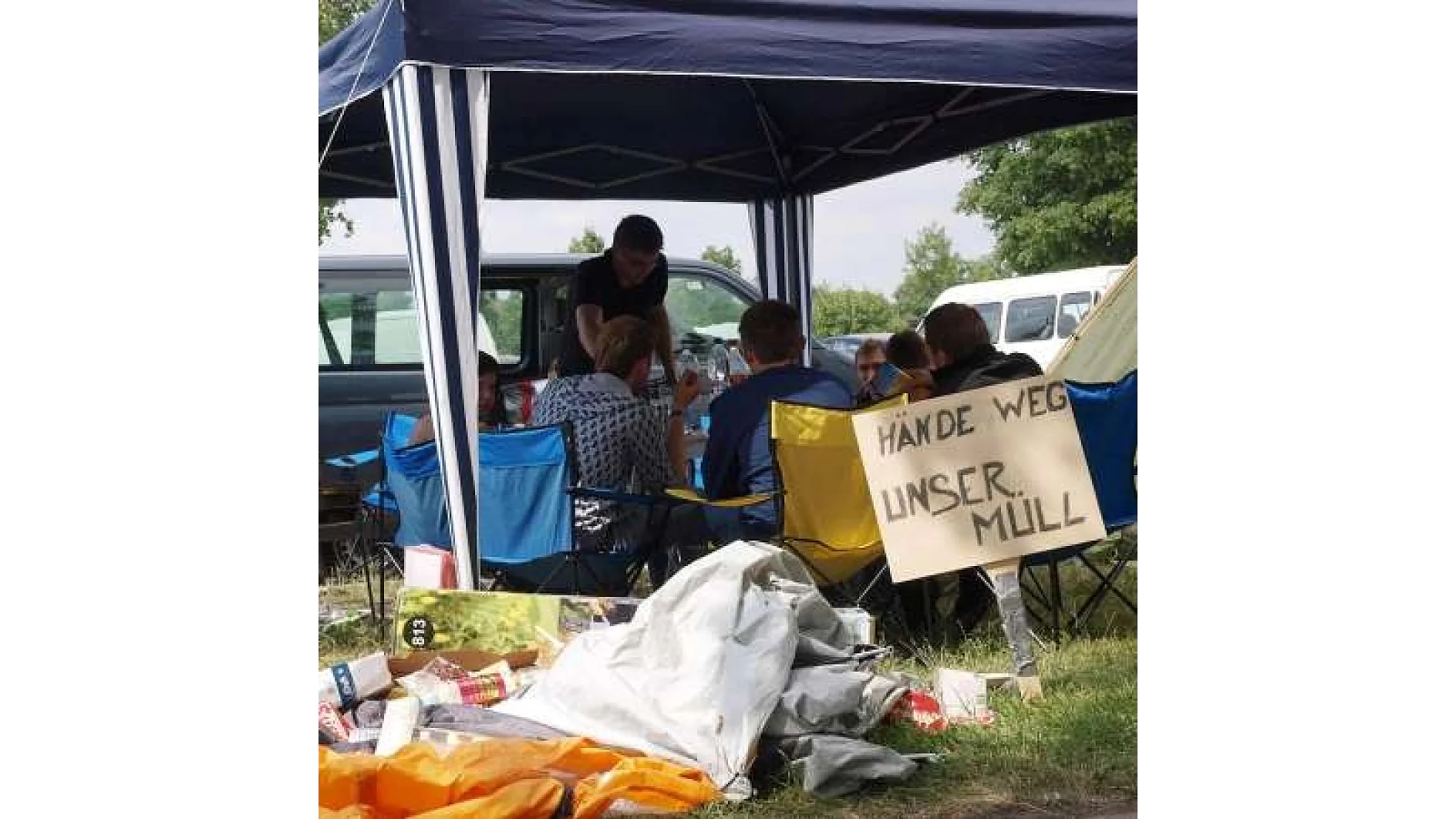 Eine Gruppe von Menschen sitzt unter einem blau-weiß gestreiften Baldachin mit verschiedenfarbigen Stühlen. Auf einem Schild steht Hände weg unser Müll. Campingausrüstung und mit Tesa-Klebeband befestigte Gegenstände liegen verstreut auf dem Gras in der Nähe. (This text has been generated by AI)