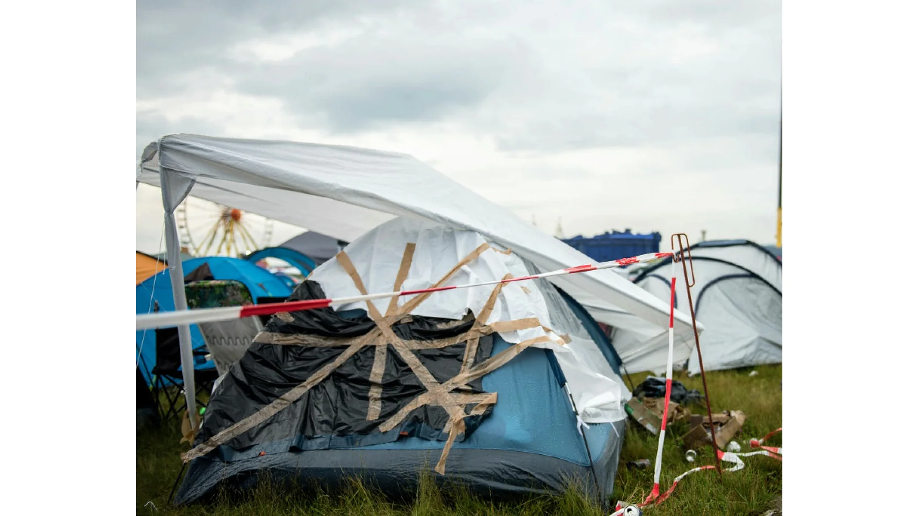 Ein provisorisches Zelt ist mit Tesa-Klebeband geflickt und mit einer Plane abgedeckt. Es steht auf Gras, in der Nähe stehen weitere Zelte. Im Hintergrund ist ein Riesenrad unter einem bewölkten Himmel zu sehen. An einer Seite ist rotes und weißes Tesa-Klebeband befestigt. (This text has been generated by AI)