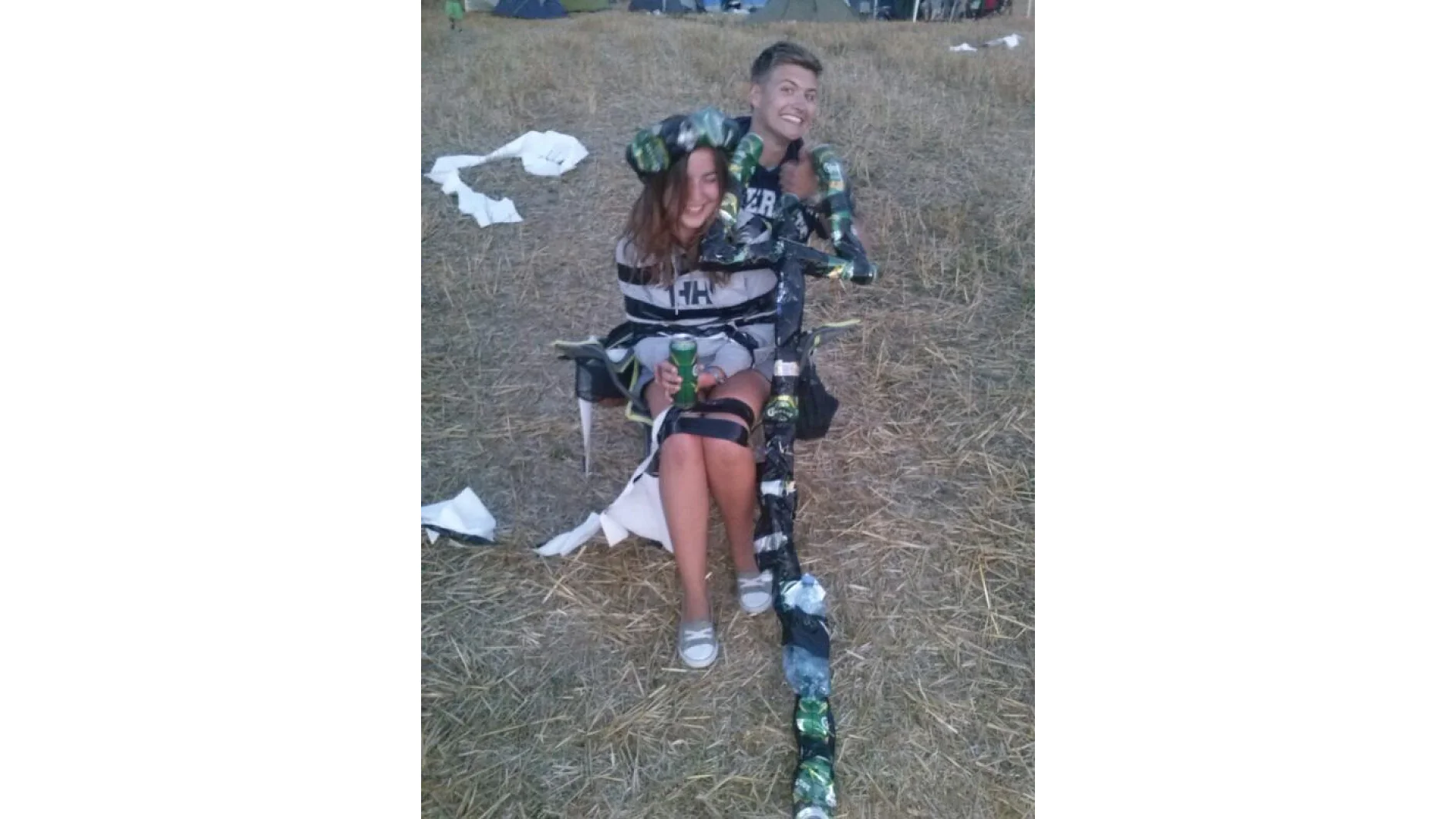 Zwei Menschen stehen draußen auf einem Feld mit trockenem Gras. Einer sitzt auf einem Stuhl, ist mit Tesa-Klebeband und leeren Dosen umwickelt und hält ein Getränk in der Hand. Der andere steht dahinter, lächelt und gestikuliert mit dem Daumen nach oben. Zerrissenes Papier liegt verstreut auf dem Boden. (This text has been generated by AI)