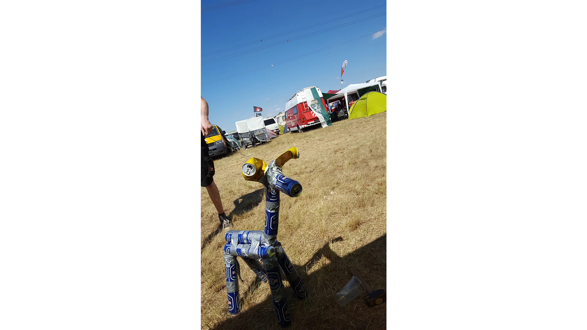 Eine Skulptur aus leeren Bierdosen, die einem Hund ähnelt, steht auf trockenem Gras. Im Hintergrund sind geparkte Fahrzeuge, Zelte und ein blauer Himmel zu sehen. Links ist teilweise der Arm einer Person zu sehen. (This text has been generated by AI)