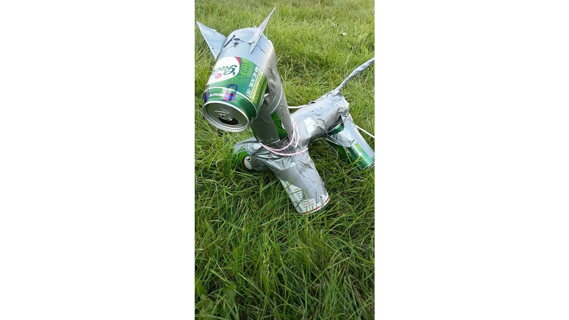 Aus leeren Getränkedosen und silbernem Tesaband ist eine Skulptur in Form eines kleinen Tieres entstanden. Es steht auf Grasboden und hat vier Beine, einen Schwanz und einen Kopf aus den Dosen. (This text has been generated by AI)