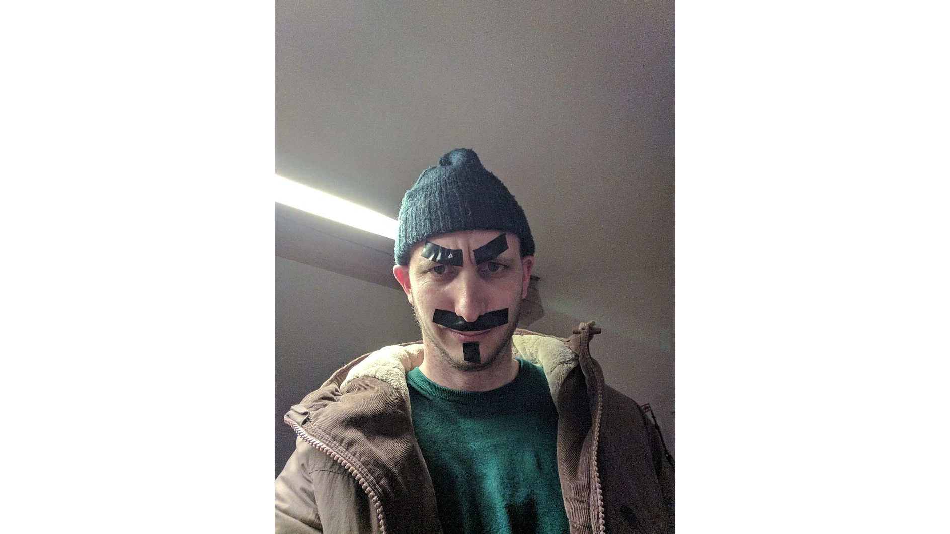 Eine Person mit dunkler Beanie und brauner Jacke hat schwarzes Tesa-Klebeband im Gesicht, das so angeordnet ist, dass es Augenbrauen, einem Schnurrbart und einem Spitzbart ähnelt. Sie befindet sich in einem Raum mit einem hellen Licht im Hintergrund. (This text has been generated by AI)