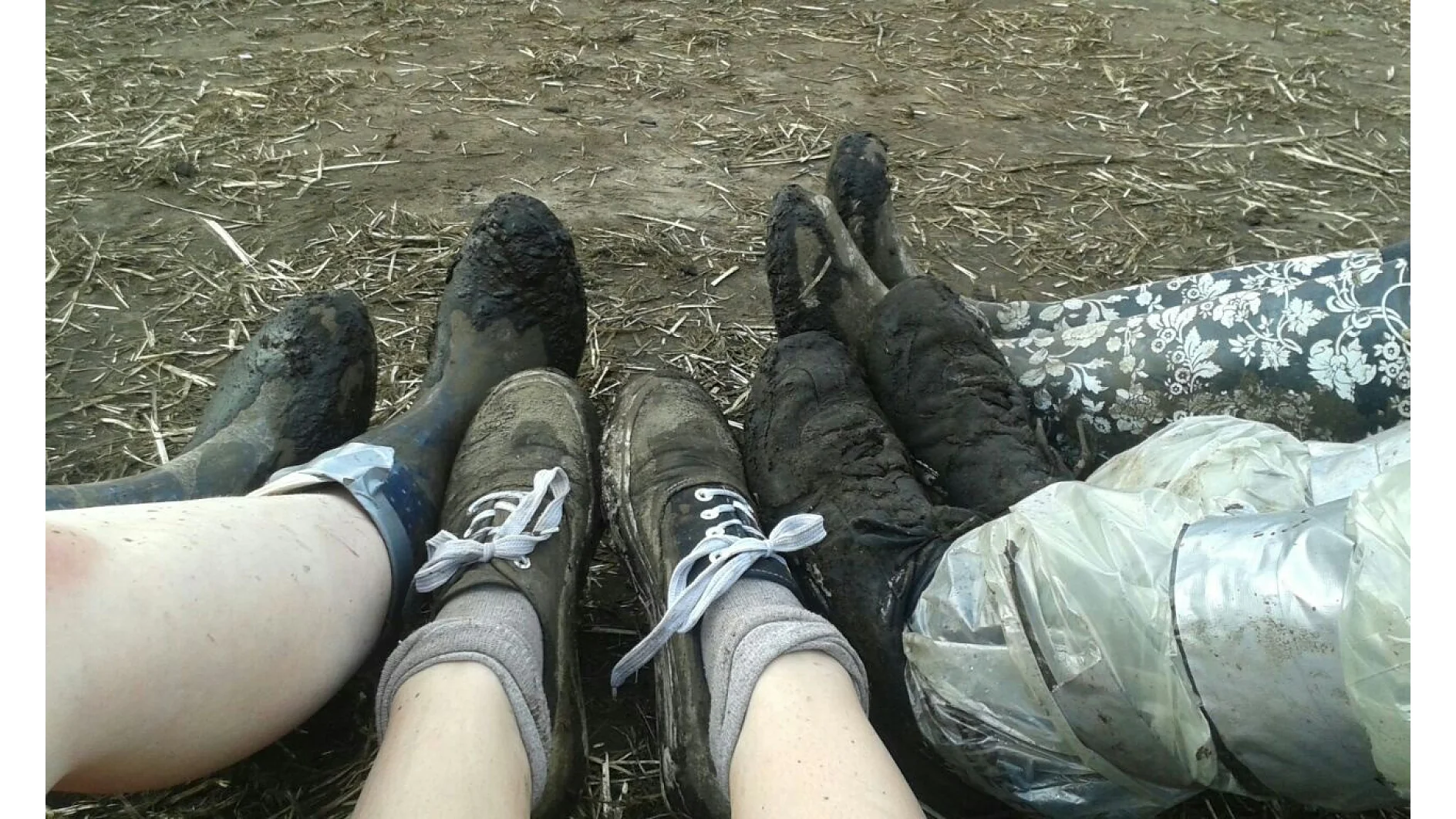 Vier Paar schlammbedeckte Stiefel und Beine liegen auf einem Erdboden. Die Stiefel haben unterschiedliche Stile und die Beine sind mit unterschiedlichen Mustern versehen, darunter einfarbige und florale Muster. Einige haben durchsichtiges Plastik, das mit Tesa-Klebeband befestigt ist und Teile der Beine bedeckt. (This text has been generated by AI)
