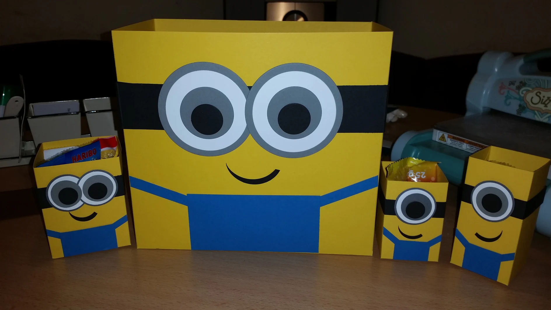 Drei gelbe Papierschachteln mit Cartoon-Minion-Motiven stehen auf einem Holztisch. Eine große Schachtel steht in der Mitte, daneben zwei kleinere. Die Schachteln haben aufgedruckte Augen, Smileys und blaue Overalls, die an Minions erinnern. (This text has been generated by AI)