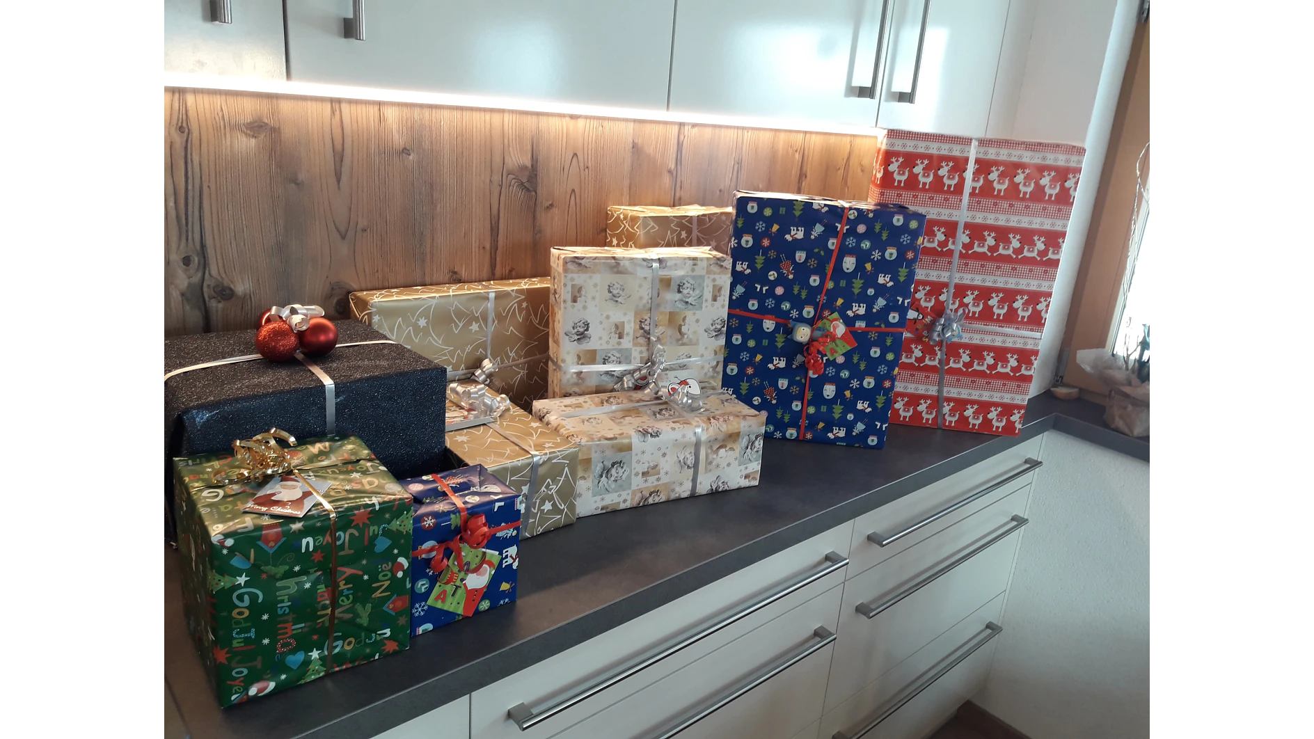 Auf einer Küchentheke stehen mehrere verpackte Geschenke. Die Geschenke sind in verschiedenen Größen und Farben erhältlich, darunter Gold, Blau mit Schneeflocken und Rot mit Rentiermuster. Jedes Paket ist mit Tesa-Klebeband befestigt, was für ein ordentliches Finish sorgt. Einige haben dekorative Schleifen; ein paar rote Kugeln liegen auf dem dunklen Paket. (This text has been generated by AI)