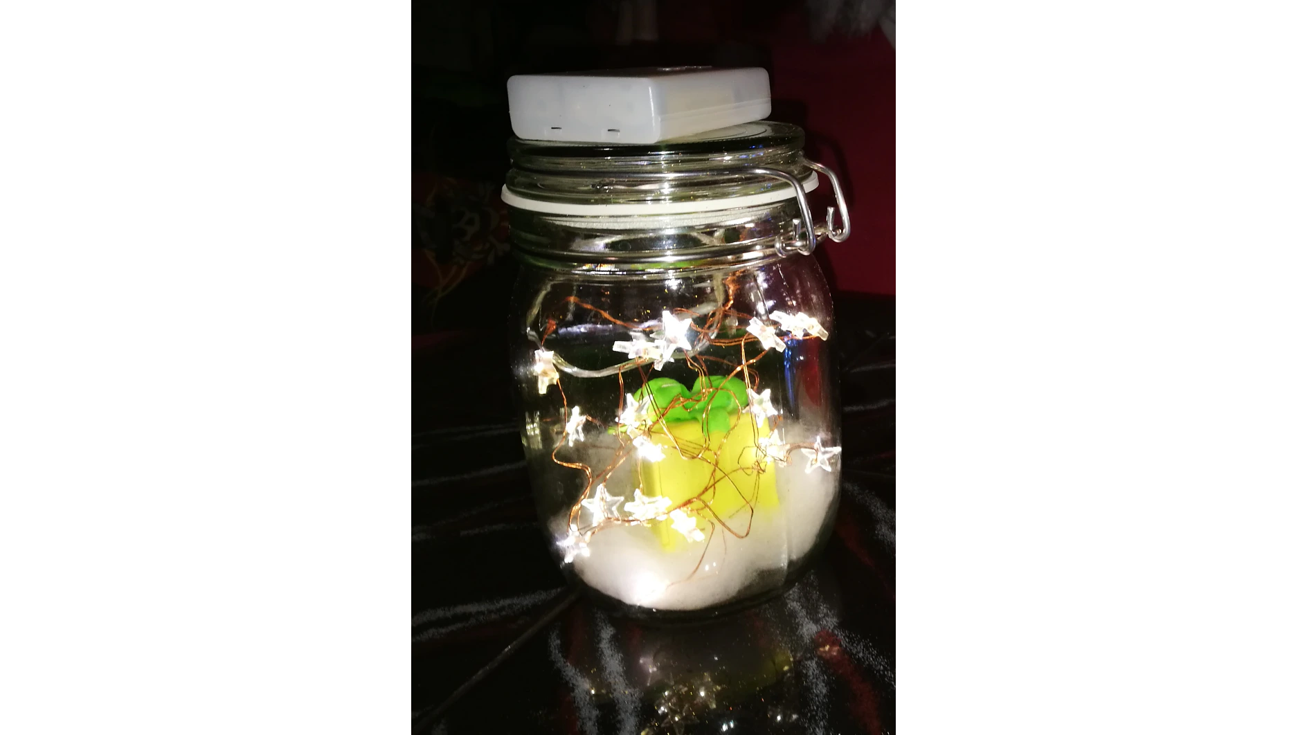 Ein Glasgefäß mit festgeklemmtem Deckel enthält eine Reihe kleiner, sternförmiger Lichter. Die Lichter leuchten. Im Inneren des Glases scheint sich am Boden ein weißes Material zu befinden und in der Mitte ein leuchtend gelbes Objekt, alles mit Tesa-Klebeband befestigt. (This text has been generated by AI)