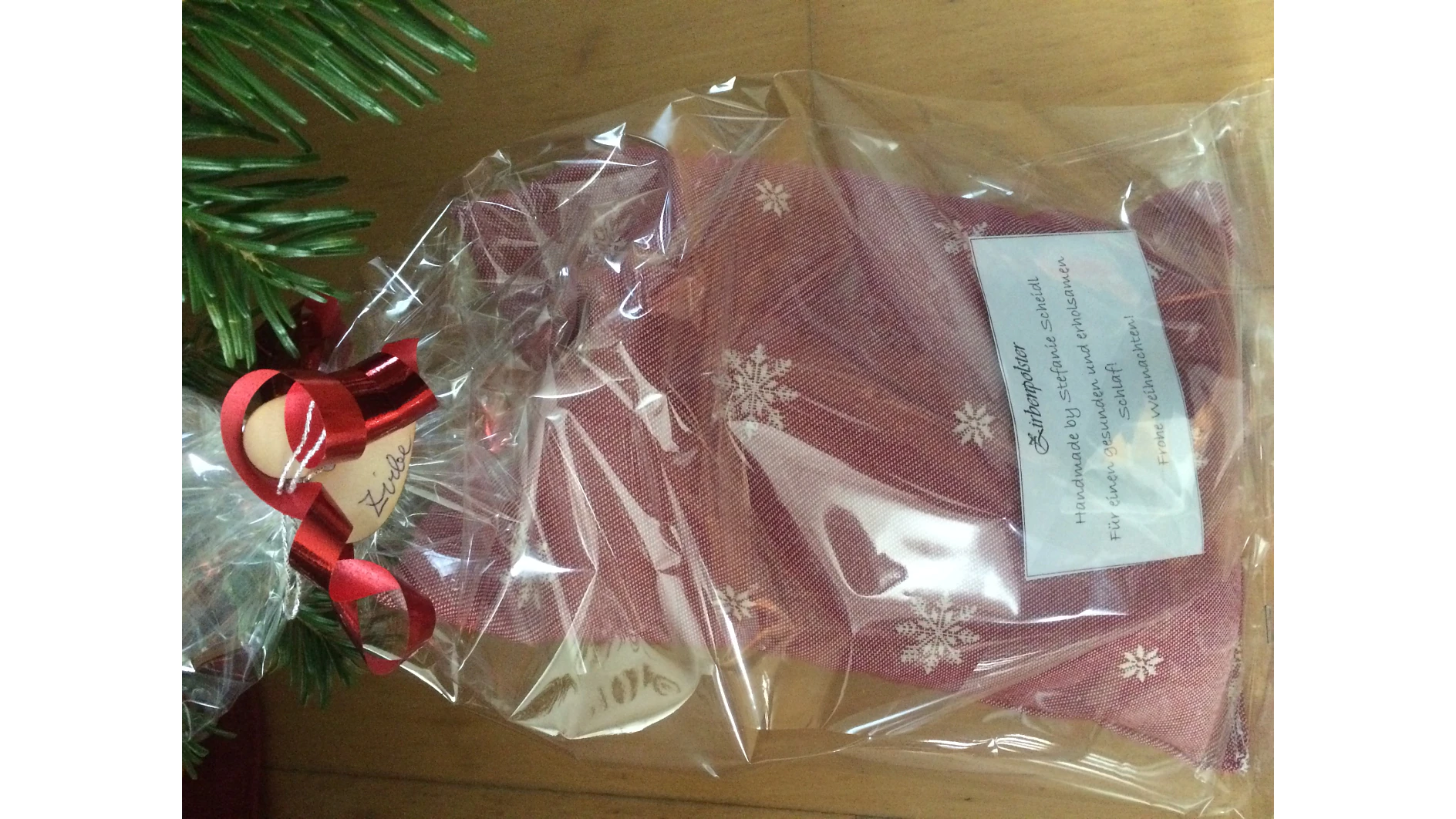 Ein roter Stoffbeutel mit weißen Schneeflocken ist in durchsichtige Plastikfolie eingewickelt und mit einem roten Band aus Tesa-Klebeband verschnürt. Ein Etikett mit teilweise verdecktem Text ist angebracht. Das Paket wird auf einer Holzoberfläche in der Nähe von grünem Laub platziert. (This text has been generated by AI)