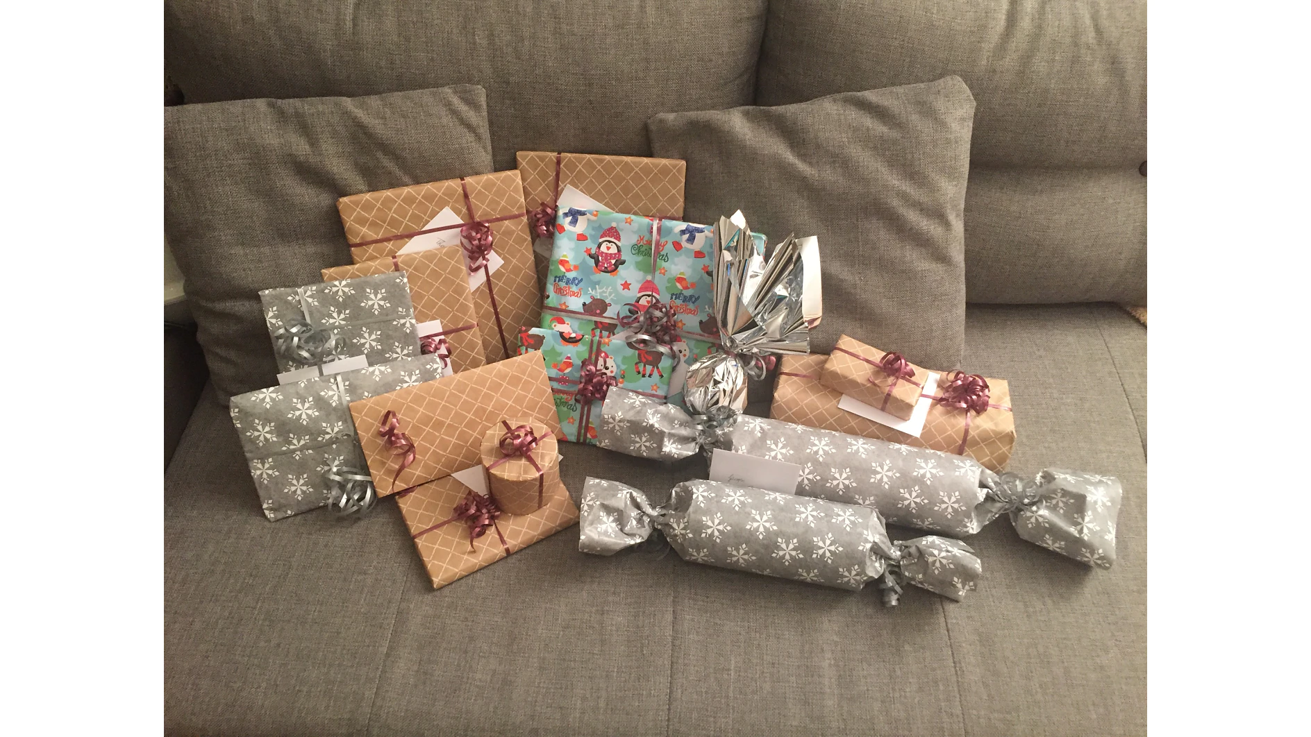 Eine Sammlung verpackter Geschenke auf einem grauen Sofa. Die Geschenke haben verschiedene Formen und Größen und sind mit braunem, silbernem und buntem Geschenkpapier versehen. Einige sind mit Schleifen und Bändern verziert, einige haben die Form großer Bonbons. (This text has been generated by AI)