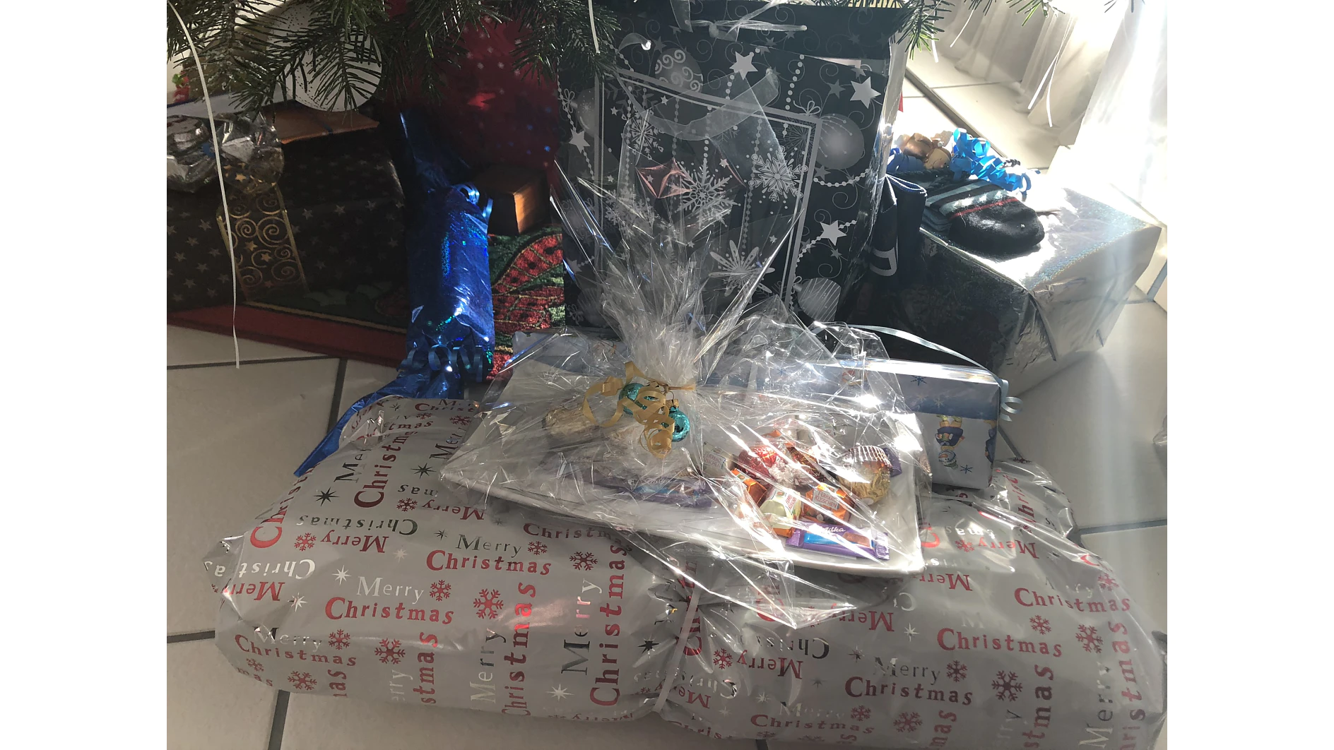 In festliches Papier eingewickelte Weihnachtsgeschenke liegen unter einem geschmückten Baum. Ein transparenter Korb, sorgfältig mit Tesa-Klebeband umwickelt, enthält obenauf verschiedene Pralinen und Leckereien. Die Szene wird durch natürliches Licht aus einem nahe gelegenen Fenster beleuchtet. (This text has been generated by AI)