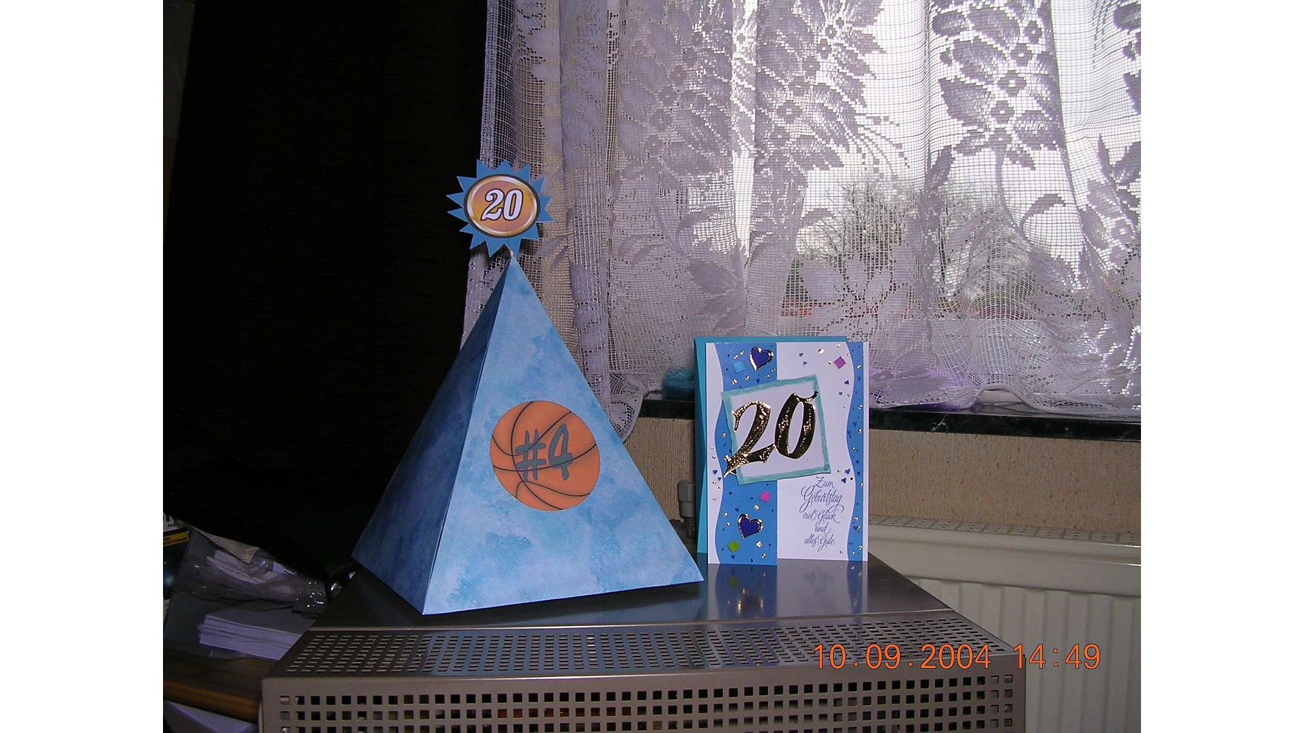 Eine blaue dreieckige Pyramide mit Basketballmotiv und einer „20“-Spitze steht neben einer blau-weißen Karte mit einer „20“ darauf. Beide stehen auf einem Tisch mit Spitzenvorhängen und einem Fenster im Hintergrund. Auf dem Foto ist das Datum „10-09-2004“ zu sehen und alles ist mit Tesa-Klebeband sicher zusammengehalten. (This text has been generated by AI)