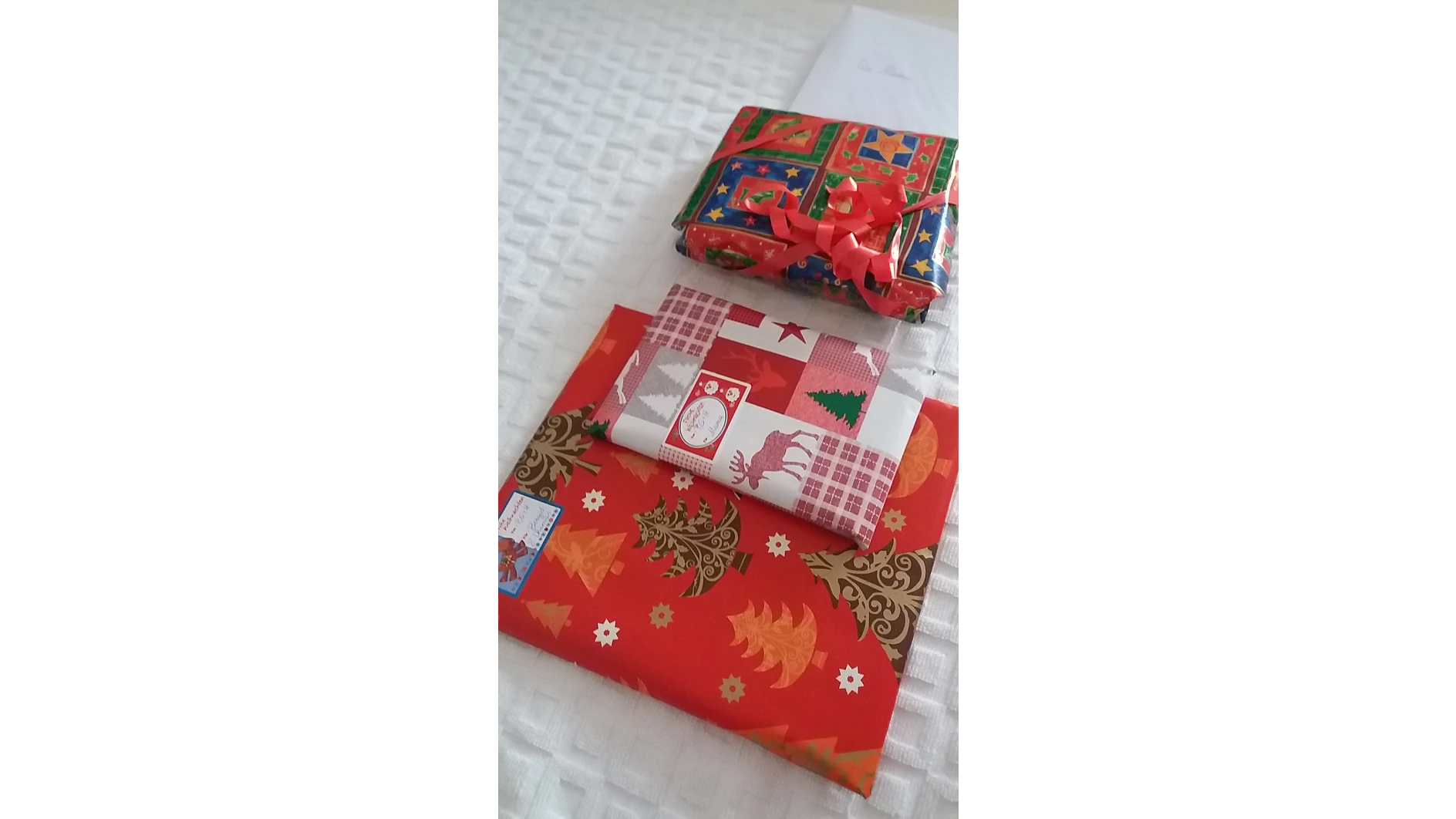 Drei verpackte Geschenke liegen auf einer weißen strukturierten Oberfläche. Zwei rechteckige Geschenke, die mit rotem und festlich gemustertem Papier mithilfe von Tesa-Klebeband befestigt sind, liegen gestapelt. Ein kleineres, quadratisches Geschenk mit grüner, roter und blauer Verpackung liegt obenauf und ist mit einer roten Schleife geschmückt. (This text has been generated by AI)