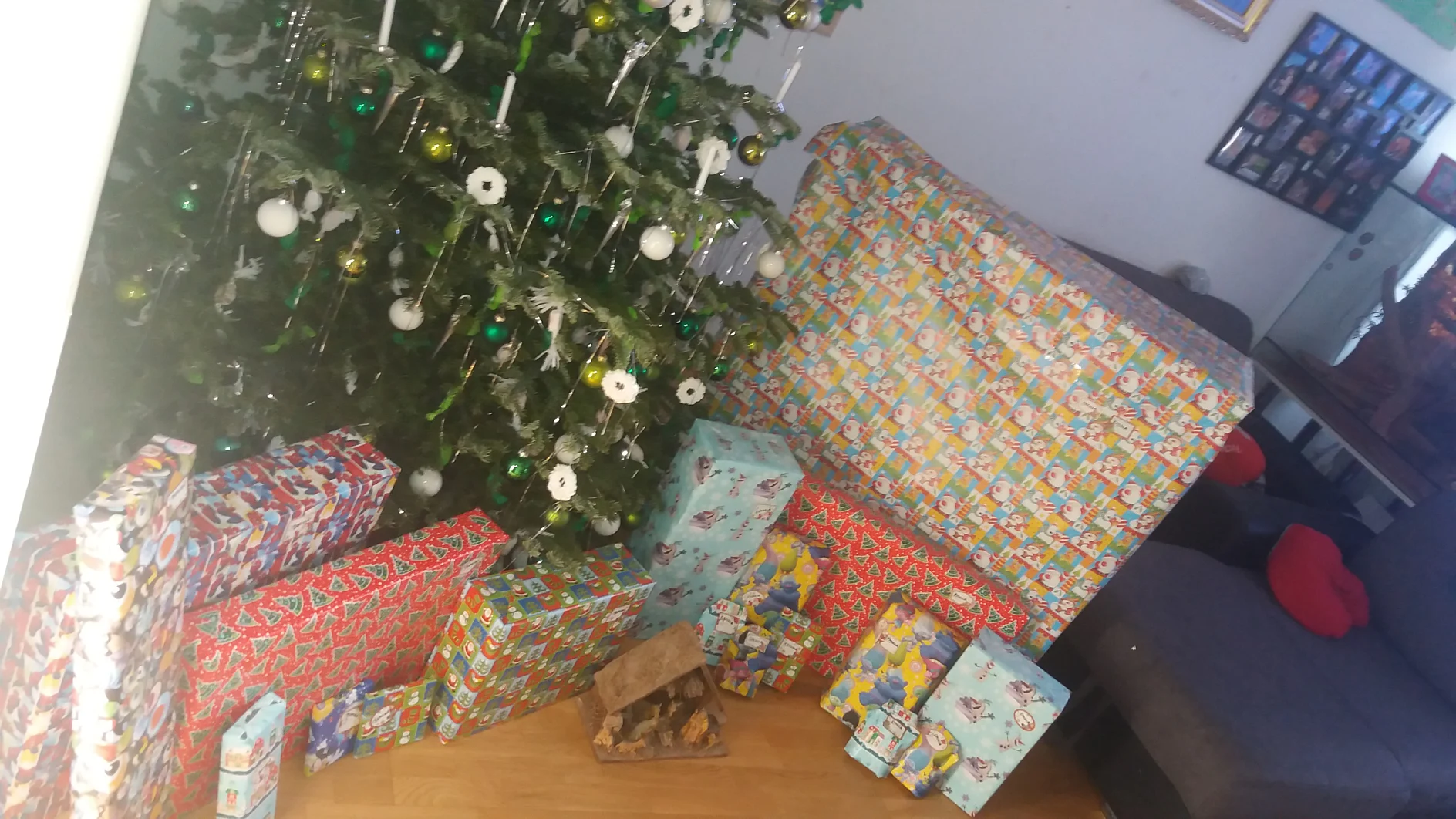 Ein geschmückter Weihnachtsbaum mit grünen und weißen Ornamenten steht neben einem großen Stapel in Weihnachtspapier verpackter Geschenke, die alle sorgfältig mit Tesa-Klebeband befestigt sind. Die Geschenke sind unterschiedlich groß und auf einem Holzboden neben einem grauen Sofa angeordnet. (This text has been generated by AI)